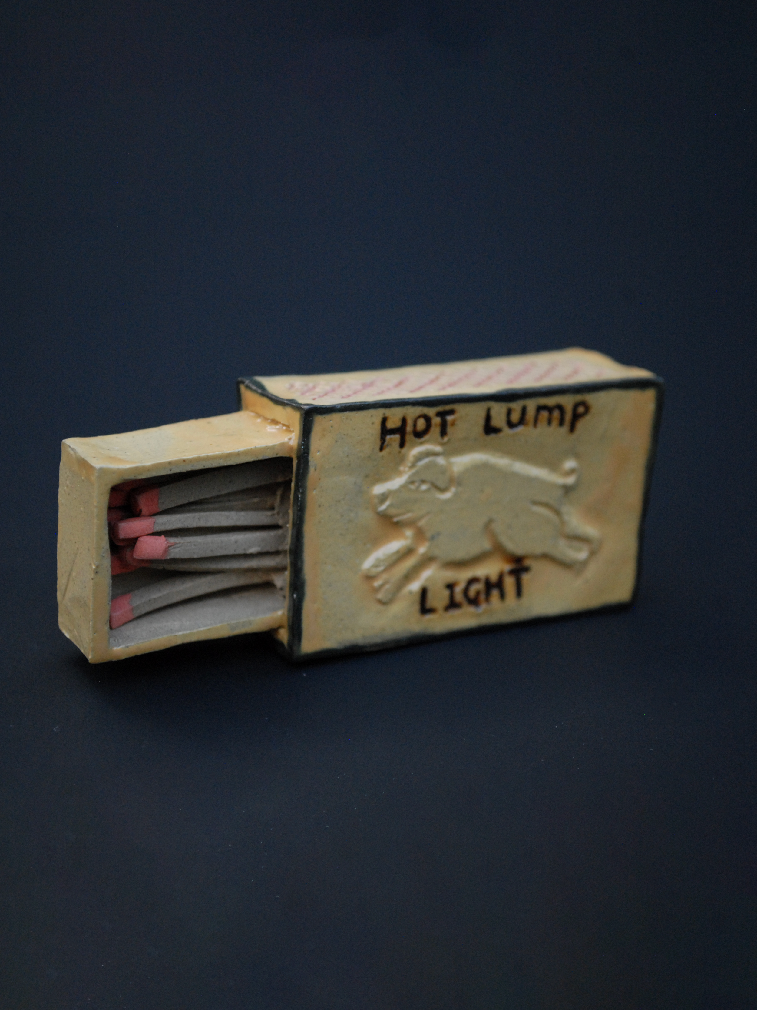 a small ceramic matchbox, with a hand-carved pig, using warm earthy tones, the words 'hot lump' are above the pig, and 'light' sits under the pig
