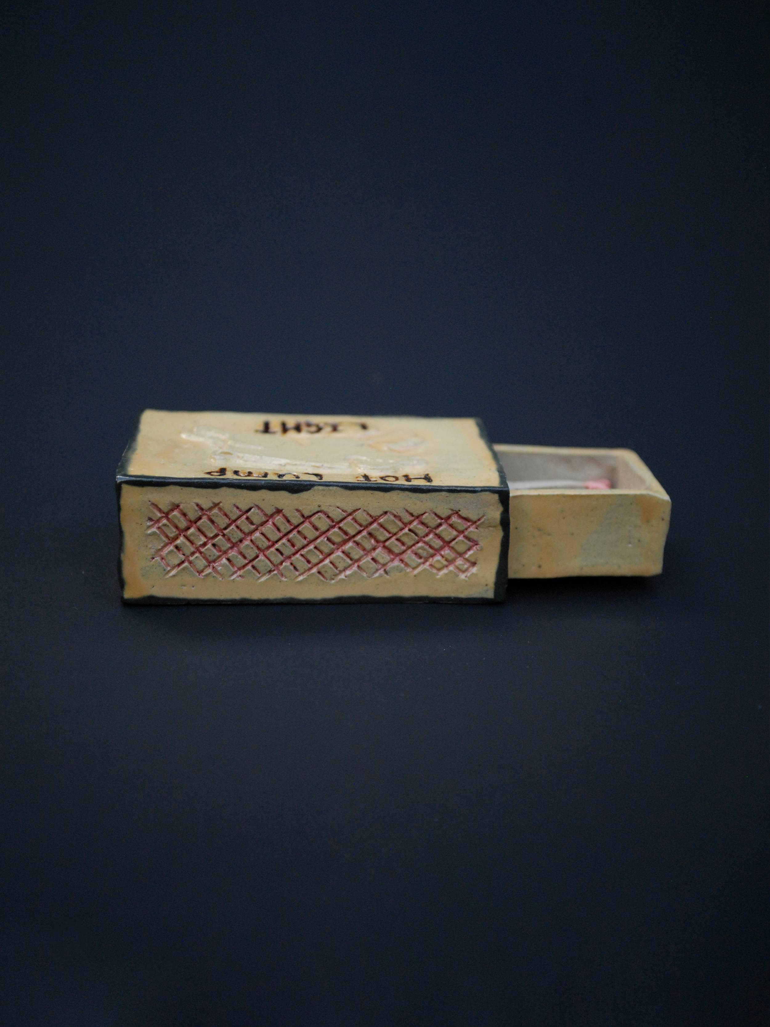 a small ceramic matchbox, with a hand-carved pig, using warm earthy tones, the words 'hot lump' are above the pig, and 'light' sits under the pig