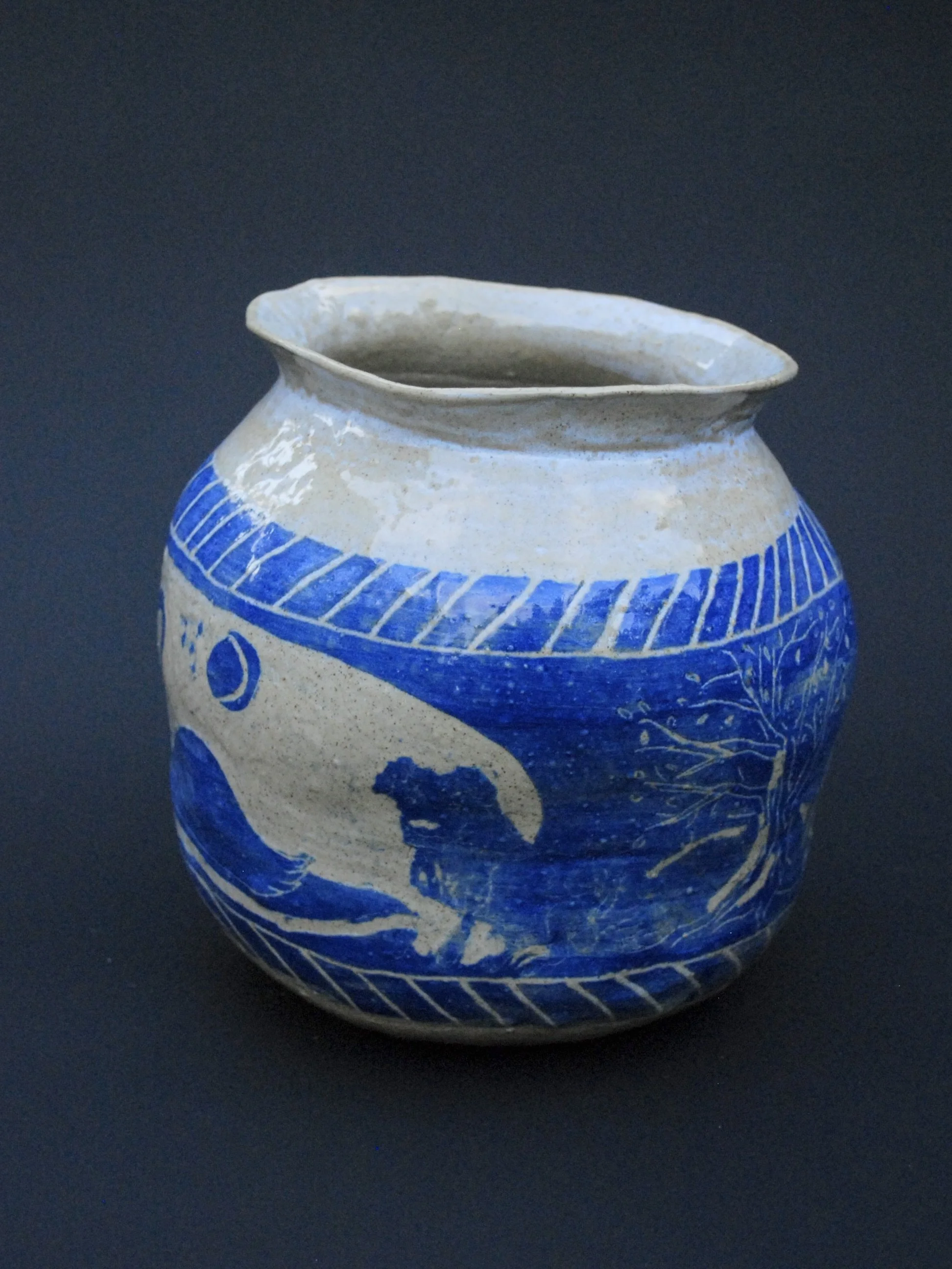 A greek inspired stoneware pot with hand-etched blue designs against a warm, earthy tone. It has a unique and slightly irregular shape adding to the rustic appeal