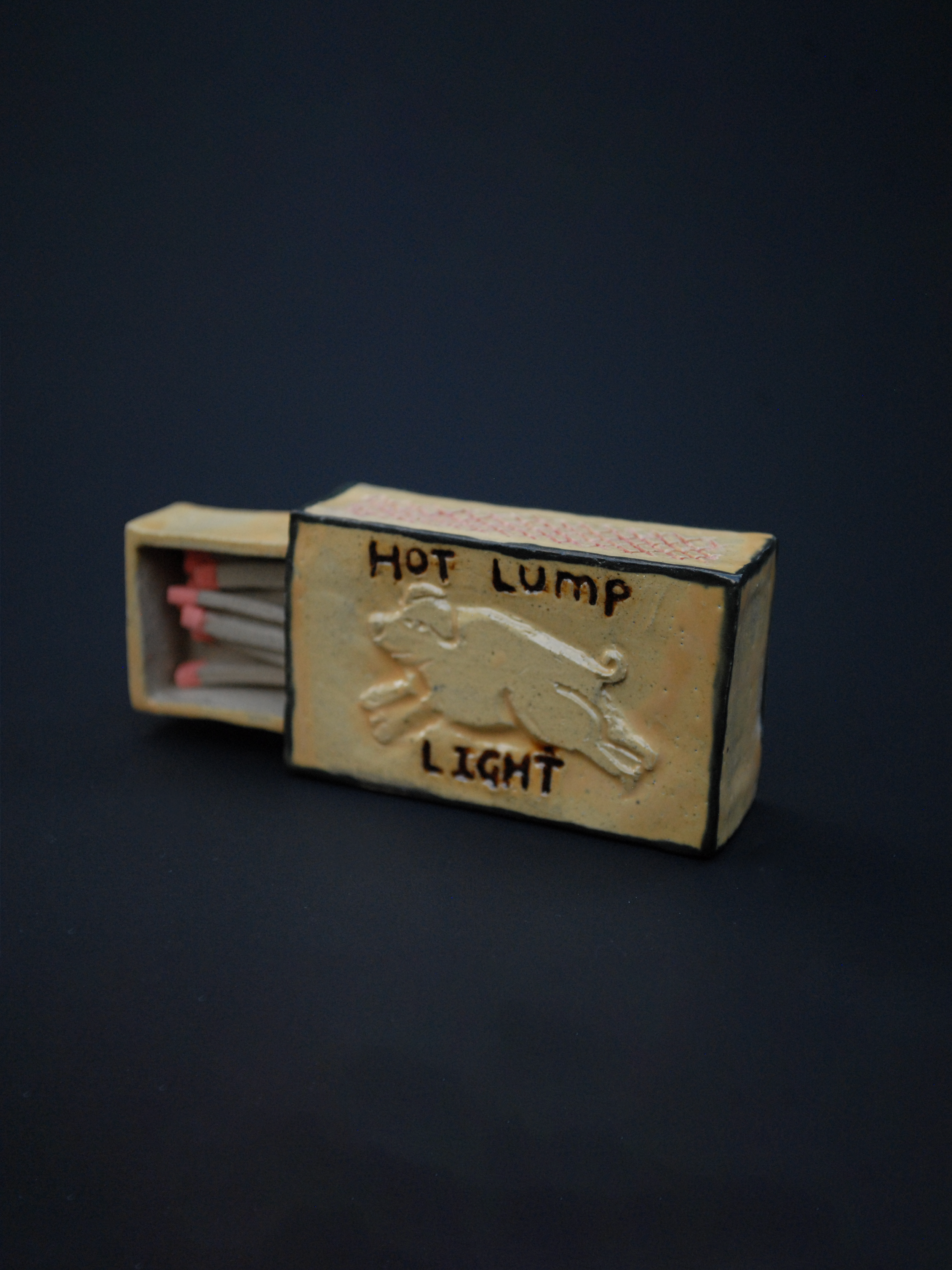 a small ceramic matchbox, with a hand-carved pig, using warm earthy tones, the words 'hot lump' are above the pig, and 'light' sits under the pig