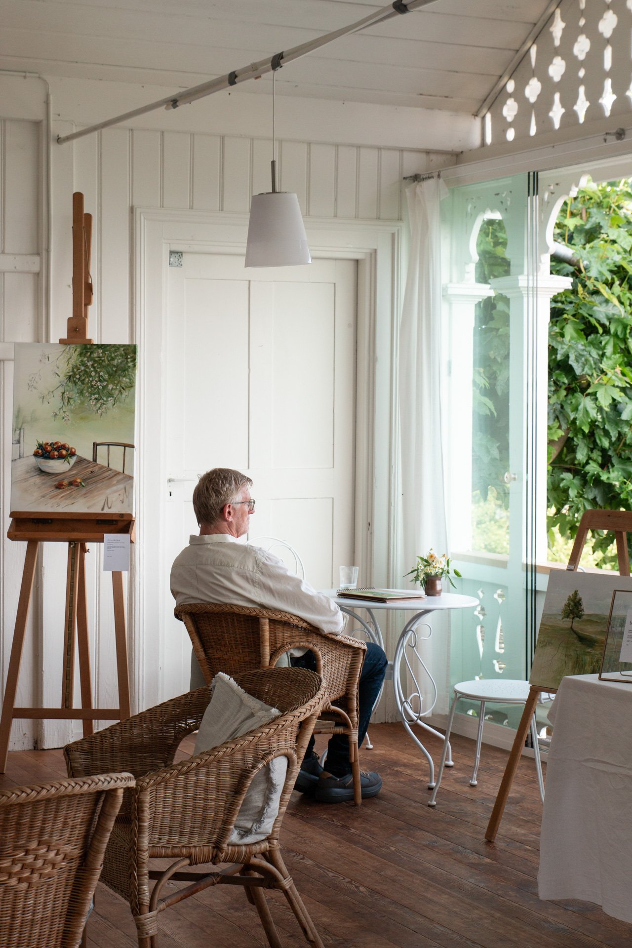Ein Mann sitzt in einem hellen, gemütlichen Raum mit weißen Wänden und Holzfußboden, neben einem offenen Fenster mit grünen Blättern draußen, und umgeben von Kunstwerken auf Ständern.