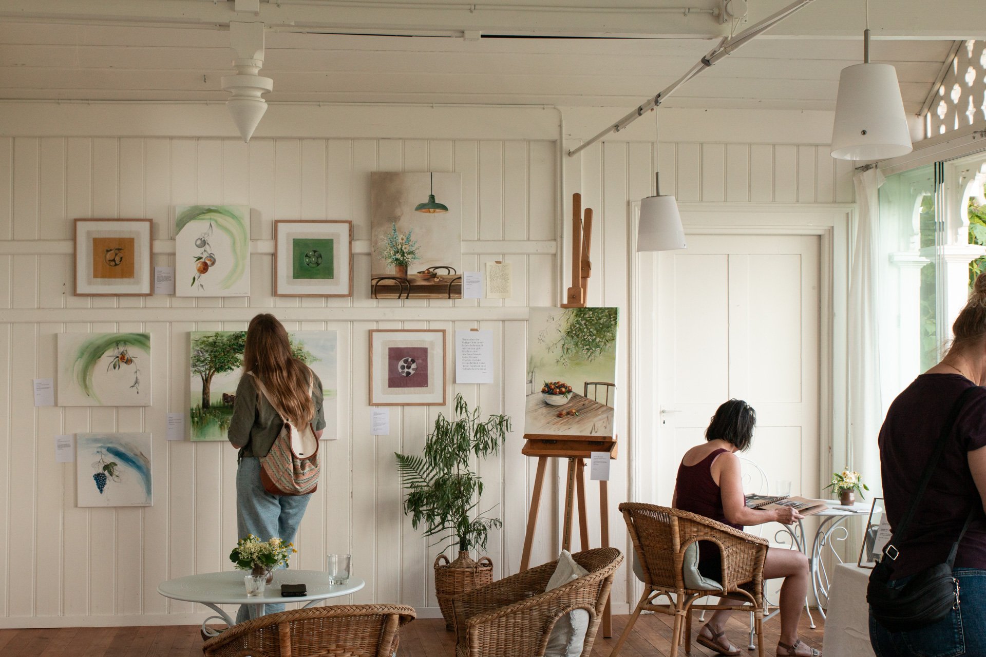 Kunstgalerie mit mehreren ausgestellten Gemälden, Besucherinnen schauen an der Wand und sitzen an Tischen, natürliche Lichtquelle durch Fenster, dekoratives Interior mit Pflanzen und Holzmöbeln.