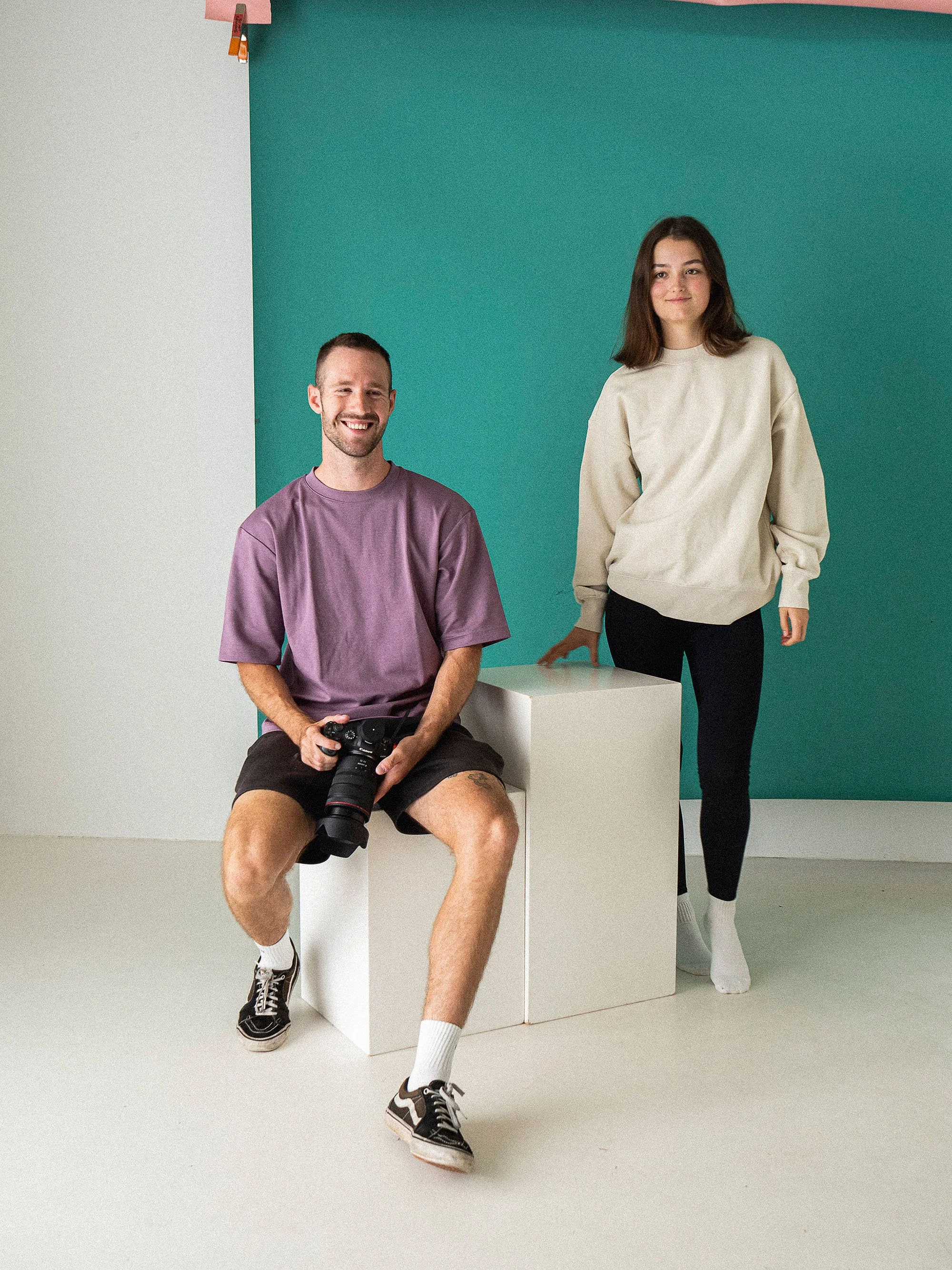 Two people in a studio with a camera, white stools, and a teal background.