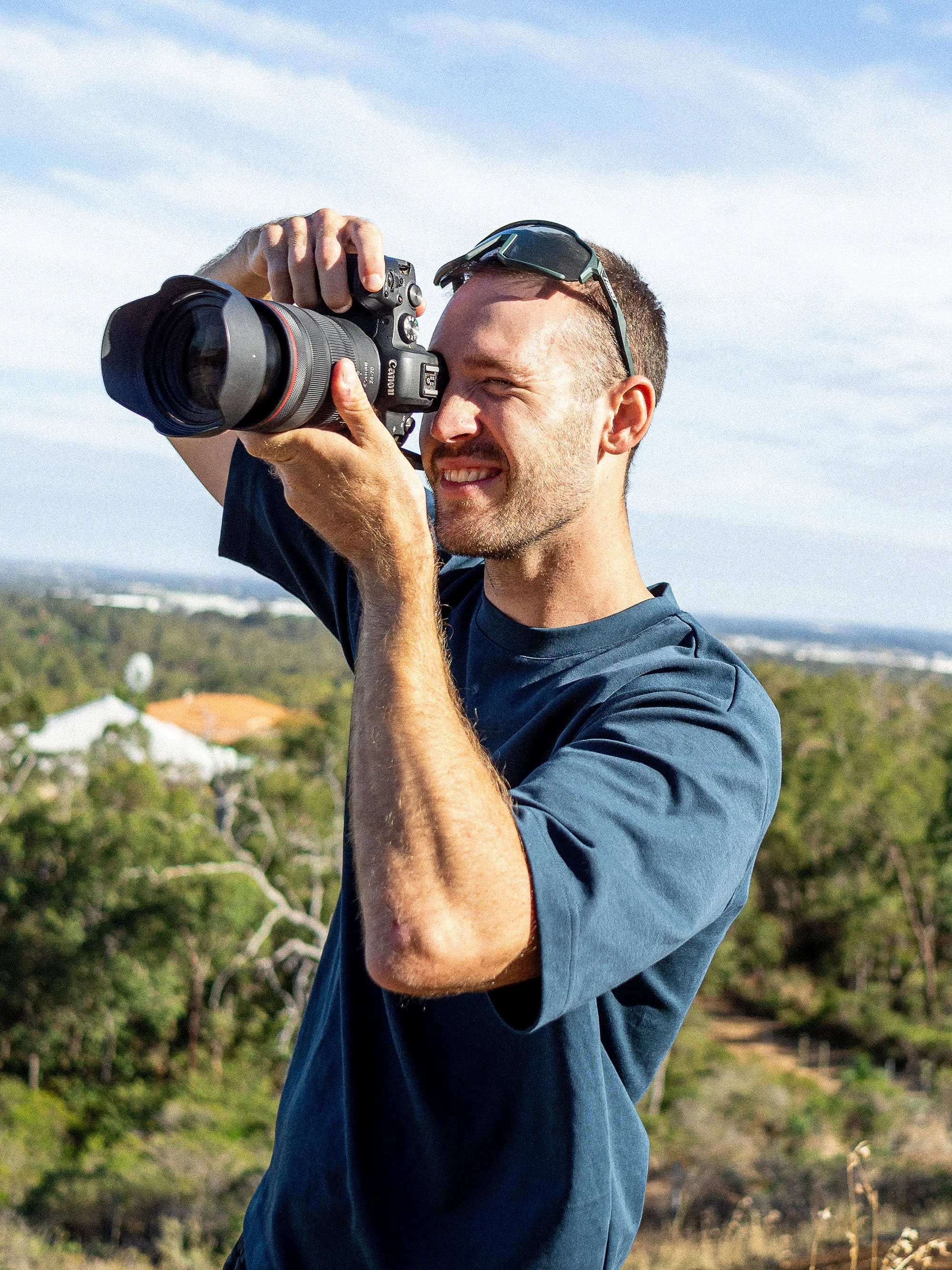Man taking a photo outdoors with a Canon camera, wearing sunglasses on his head, in a natural setting with trees and a blue sky.