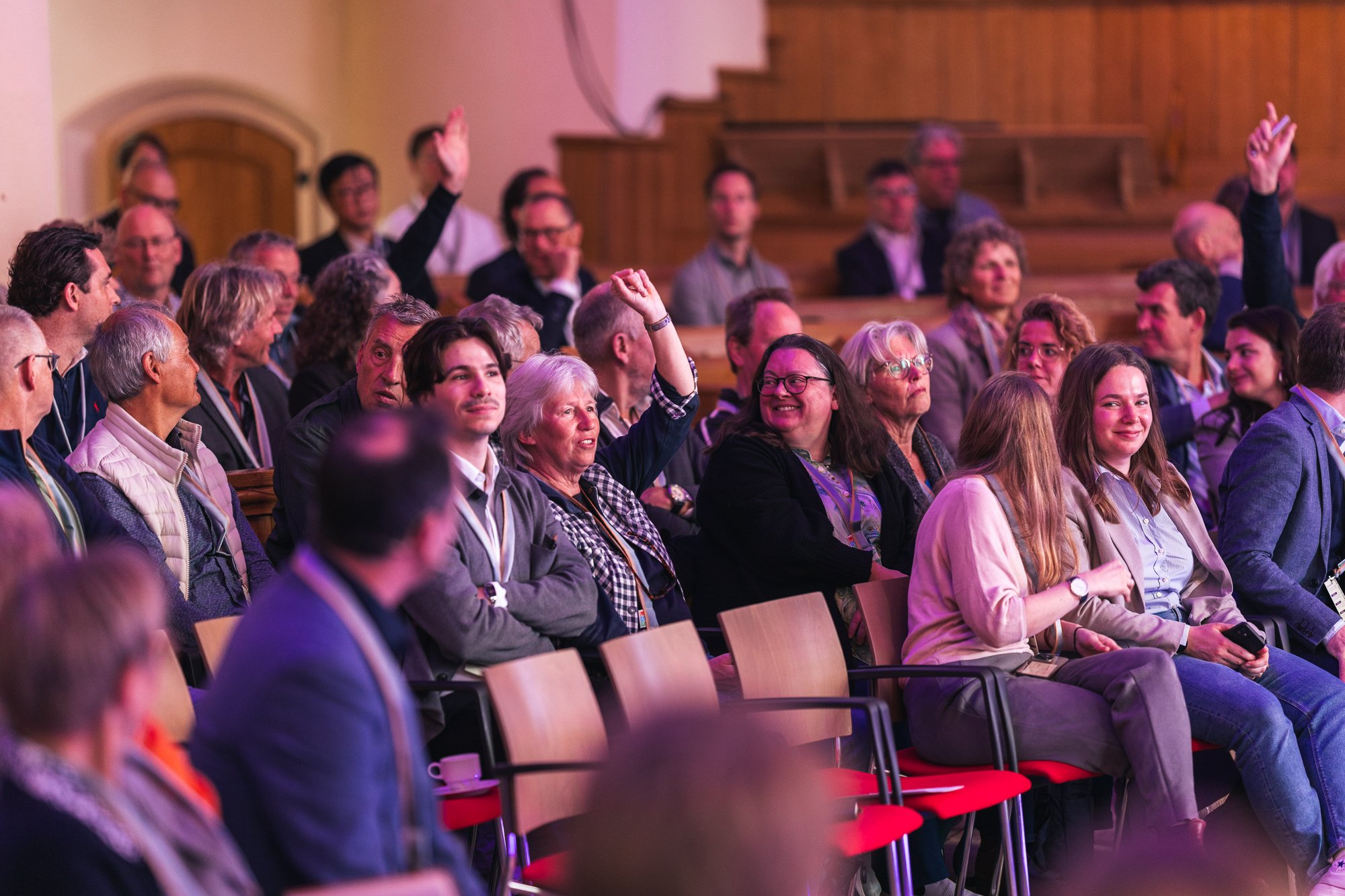 En sal fuld af mennesker, der lytter til en præsentation, med nogle få personer, der stiller spørgsmål, i en stor sal med trævægge og røde stole.