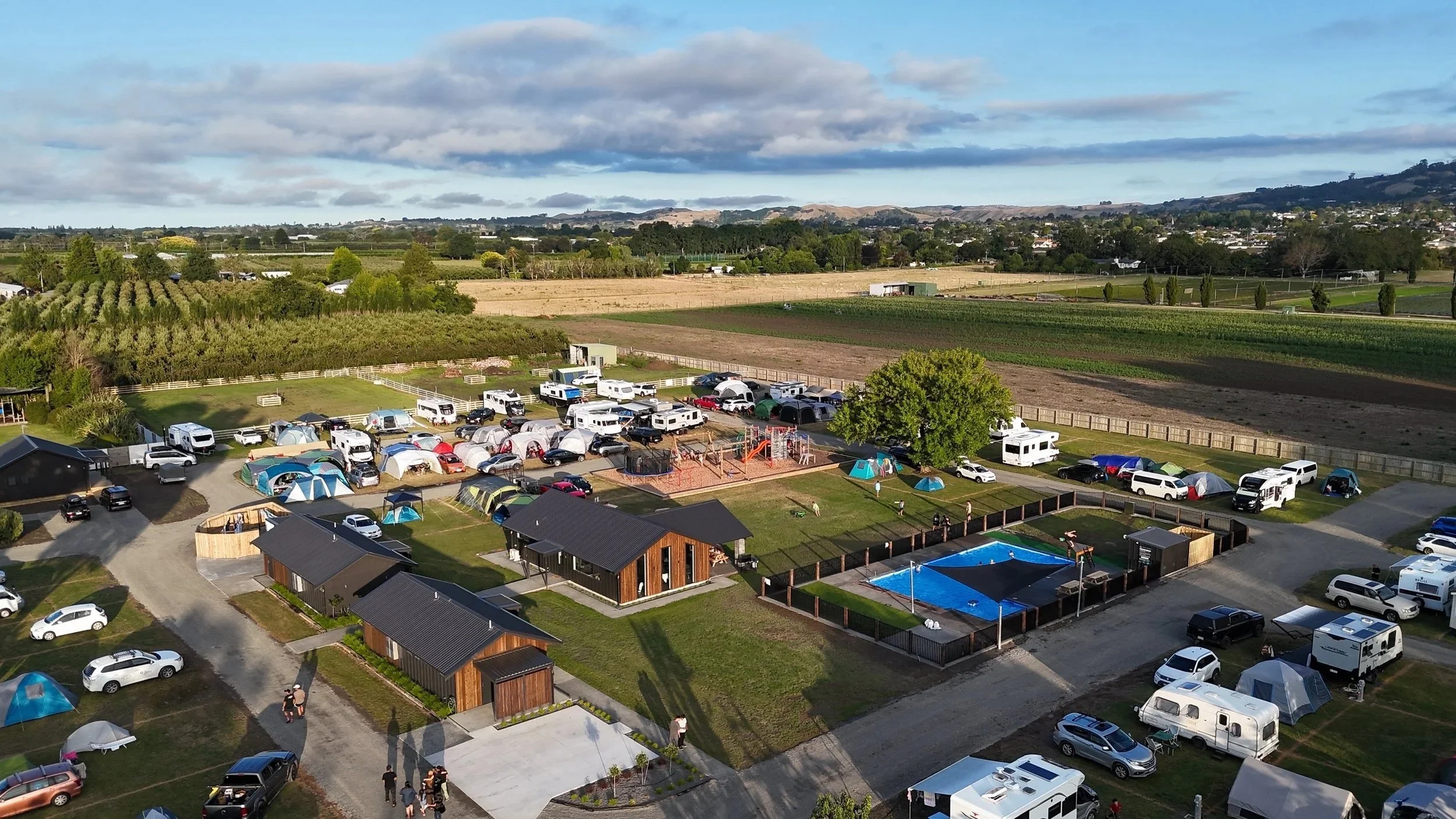 Aerial view of a campground with tents, RVs, and parked cars, a swimming pool, small buildings, and a playground, surrounded by green fields and distant hills under a partly cloudy sky.