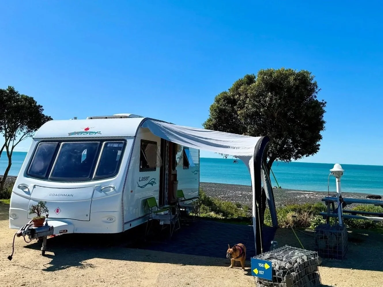 A white travel trailer with an extended canopy set up near a beach, with trees, a lamppost, and a view of the ocean in the background. A small dog is standing near the trailer, and there are some outdoor chairs, a crate, and a potted plant nearby.