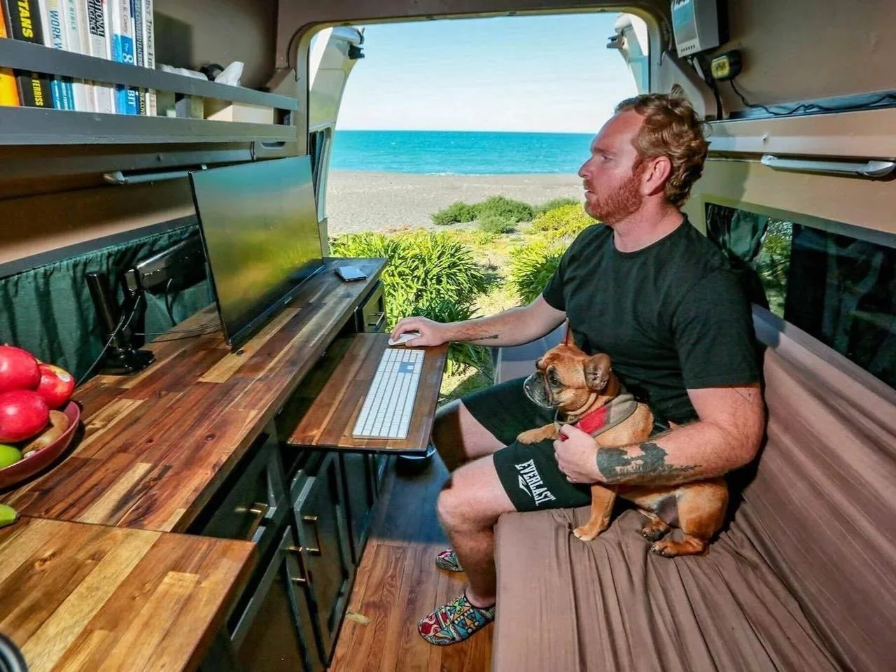 A man with red hair and beard working on a computer inside a camper van with a view of the beach and ocean outside. He is sitting on a cushioned bench with a small dog on his lap.