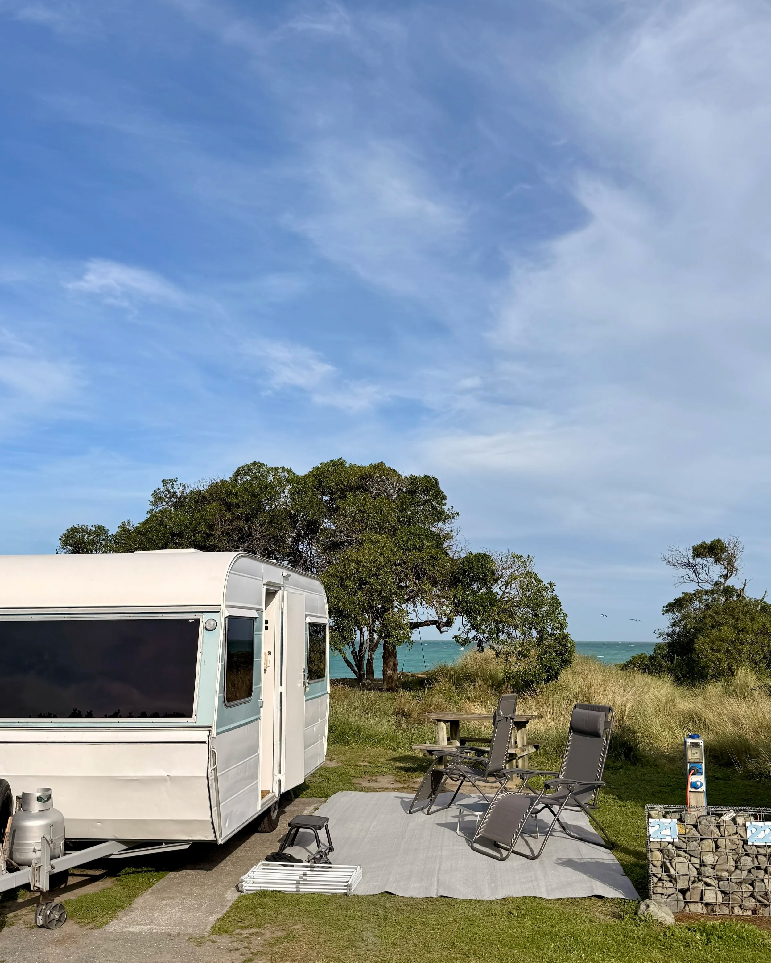 A parked camper trailer on a concrete pad near dunes with tall grass, overlooking a beach with trees in the background and a blue sky overhead.