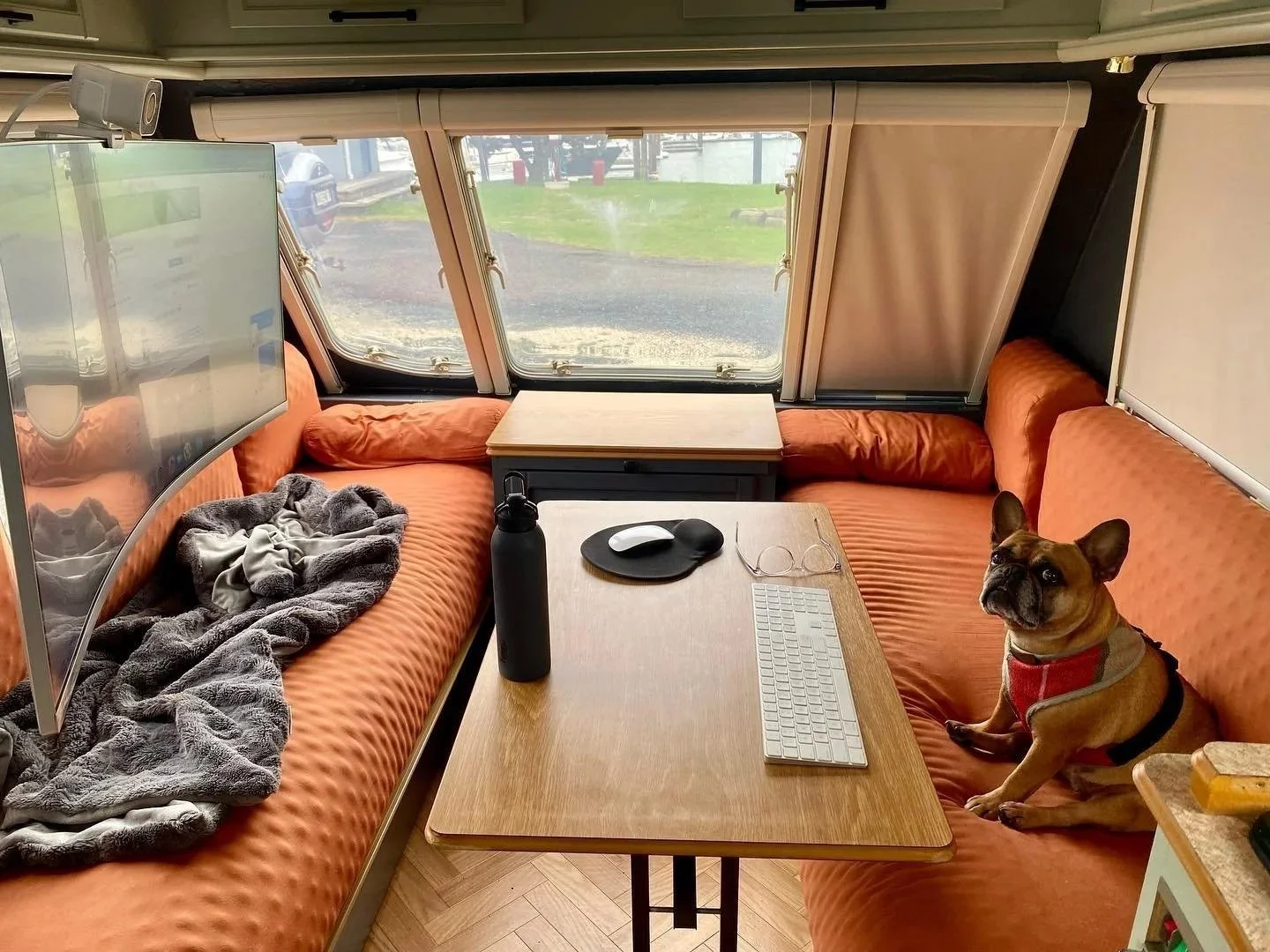 Interior of a camper van with orange cushioned seats, a small wooden table, a computer keyboard, mouse, glasses, a black water bottle, and a French bulldog wearing a red harness sitting on the seat. Outside the windows, a grassy area and parked cars are visible.
