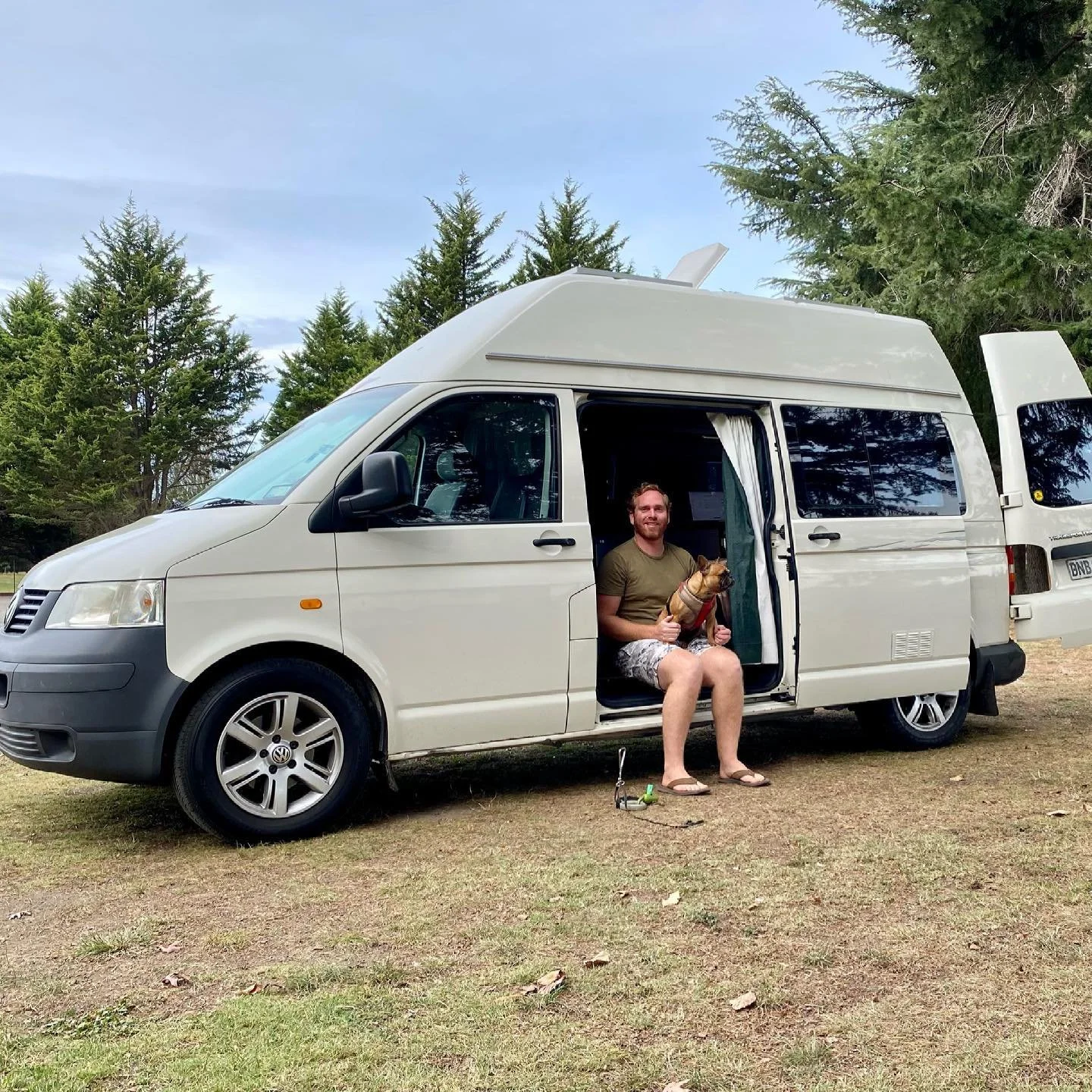 Man sitting in the doorway of a white camper van, holding a small dog, with a forested background and a cloudy sky.