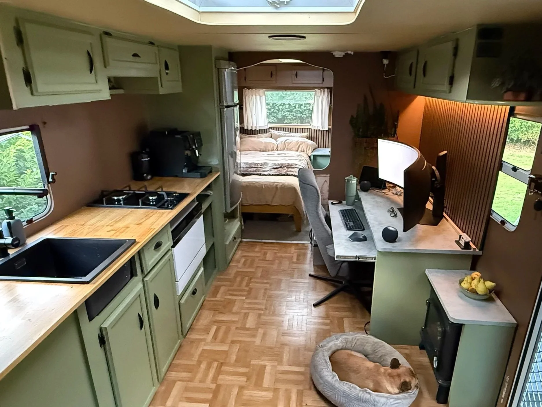 Interior of a small, cozy camper with a kitchen area on the left, a desk with a computer and monitor on the right, and a sleeping area at the back; a dog is sleeping in a bed on the floor near the desk.