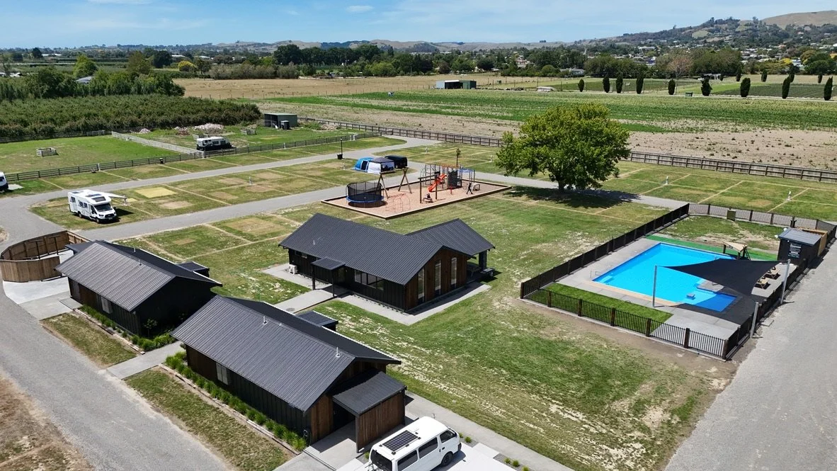 Aerial view of a recreational area with cabins, a swimming pool, a playground, and RV parking, surrounded by open fields and countryside.