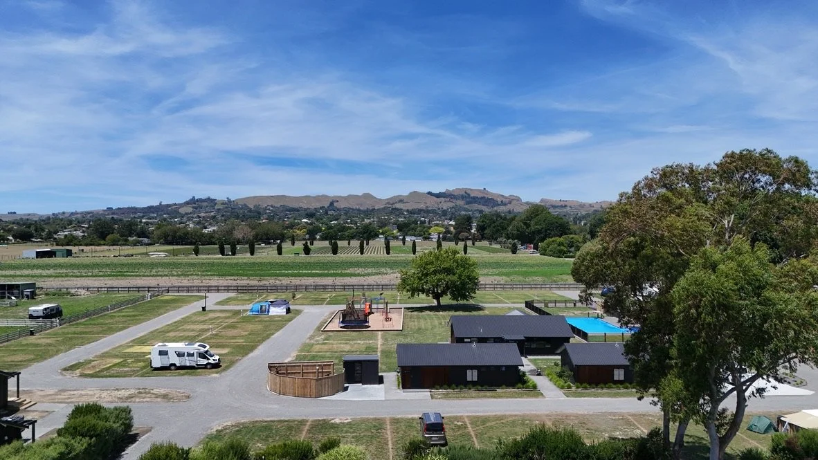 A scenic view of a campground with RVs, small cabins, a swimming pool, and a playground, surrounded by green fields, trees, and distant rolling hills under a partly cloudy sky.