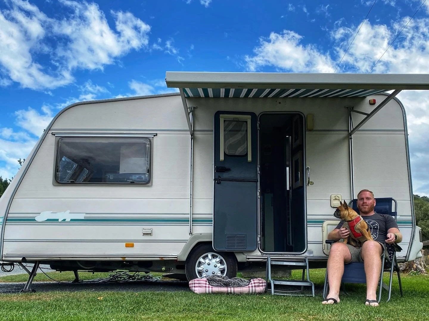 A man sitting outside a white travel trailer holding a small dog. The man is seated in a foldable chair on grass, with a dog in his lap, both looking content. The trailer has an open door, a window, and an extendable awning against a partly cloudy blue sky.