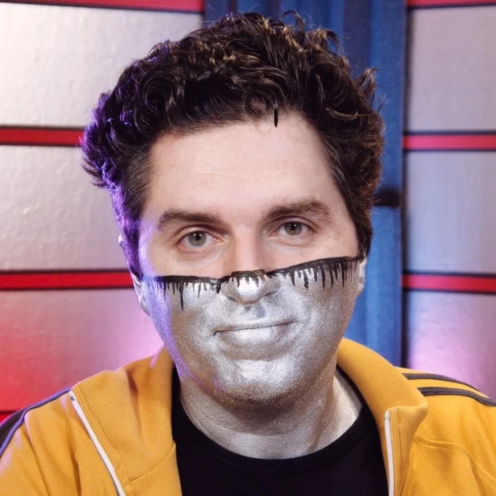 A man with dark curly hair looks directly at the camera, the lower half of his face painted silver.