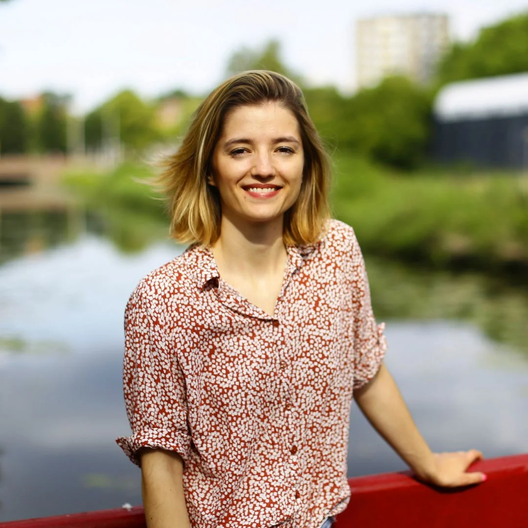 A woman with shoulder-length blonde hair smiling outdoors near a river with greenery and buildings in the background.