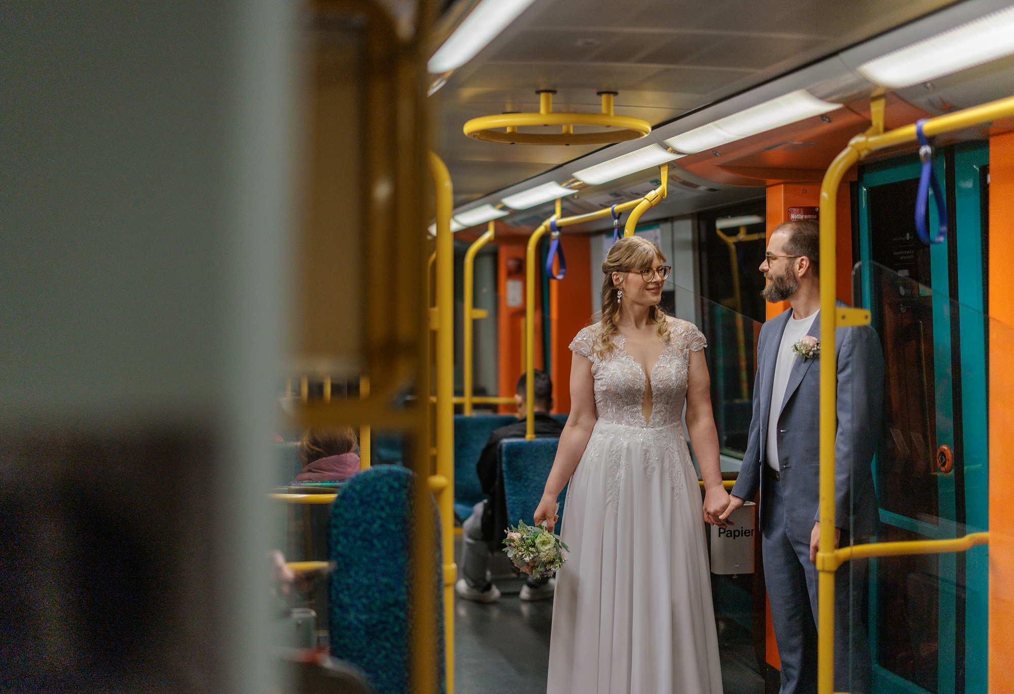 Ein Brautpaar, die Frau in einem weißen Hochzeitskleid mit Blumenstrauß, der Mann in einem grauen Anzug, hält Händchen in einem öffentlichen Verkehrsmittel, die Frankfurter U-Bahn.