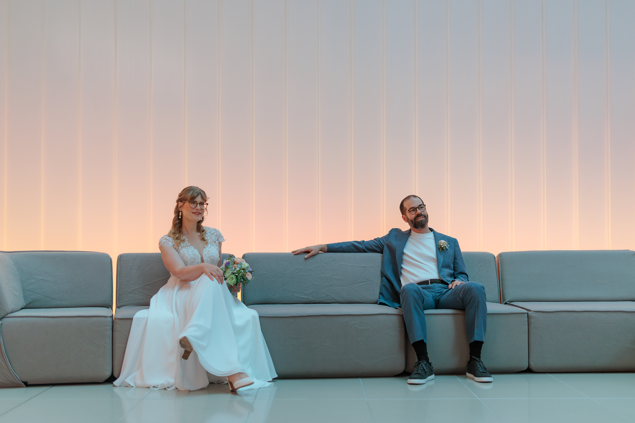Ein Brautpaar sitzt auf einem grauen Sofa in der Lobby des Eurotheum-Gebäude Frankfurt. Die Braut trägt ein weißes Hochzeitskleid und hält einen Blumenstrauß, während der Bräutigam einen dunklen Anzug trägt. Beide wirken entspannt und glücklich.