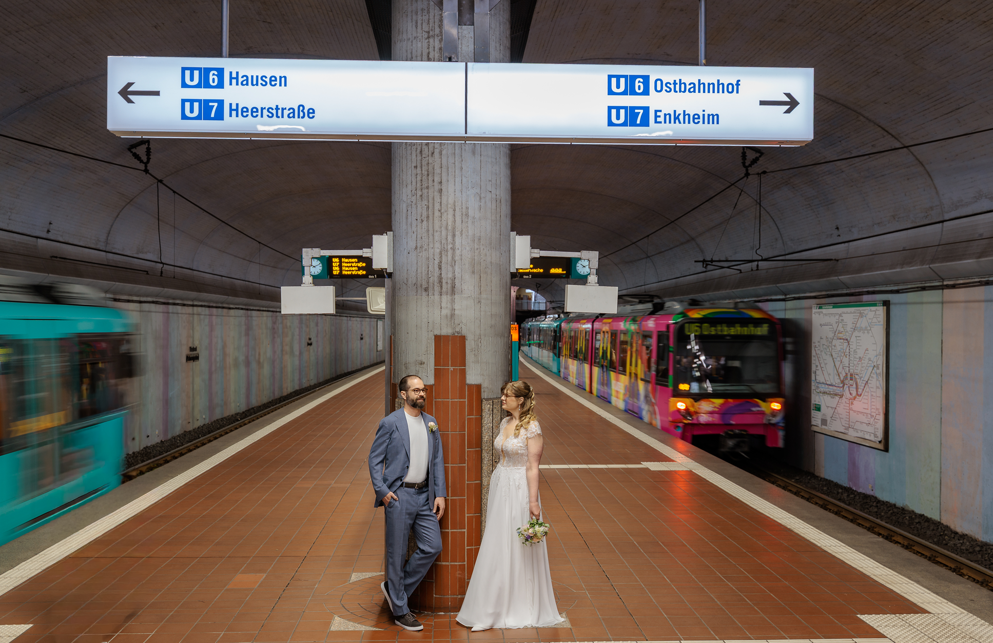 Ein Brautpaar steht in einer U-Bahn-Station, umgeben von Zügen. Die Station ist modern und hat eine große, weiße Informationsanzeige mit blauer Beschriftung, die auf die Linien U6 und U7 verweist. Das Brautpaar besteht aus einem Mann in einem hellblauen Anzug und einer Frau in einem weißen Hochzeitskleid, beide unterhalten sich. Im Hintergrund sind Bewegungsunschärfen von Zügen zu sehen.