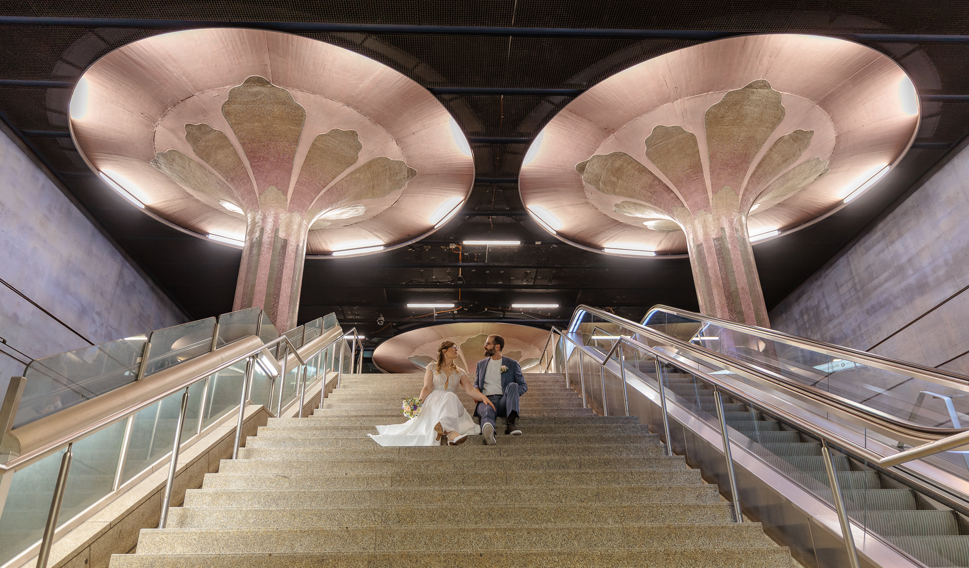 Ein Brautpaar sitzt auf den Treppen der U-Bahnstation Frankfurt Westend mit auffälligen Kupfer- oder Roségold-Decken im Hintergrund.