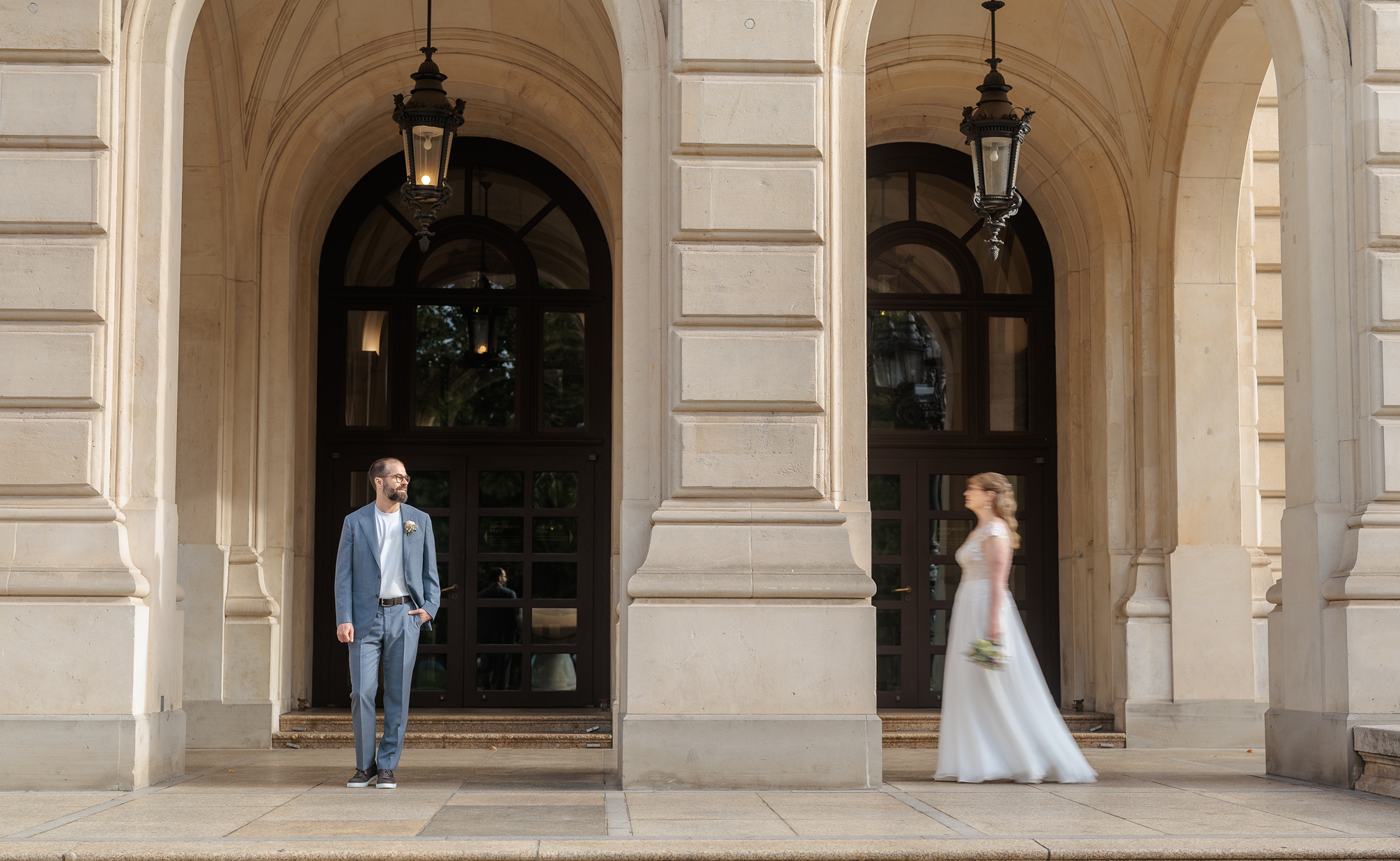 Ein Bräutigam in einem grauen Anzug und eine Frau in einem weißen Brautkleid stehen vor der Alten Oper Frankfurt. Der Mann steht rechts, schaut zur linken Seite, die Frau läuft an ihm vorbei in Richtung rechts.