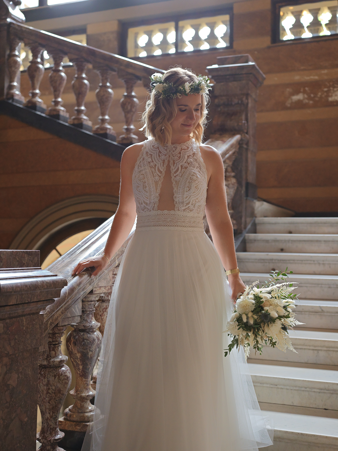 Eine junge Frau in Hochzeitskleid mit Blumenkranz auf dem Kopf steht auf einer Treppe im klassischen Gebäude, hält einen Blumenstrauß in der Hand und sieht nach unten.