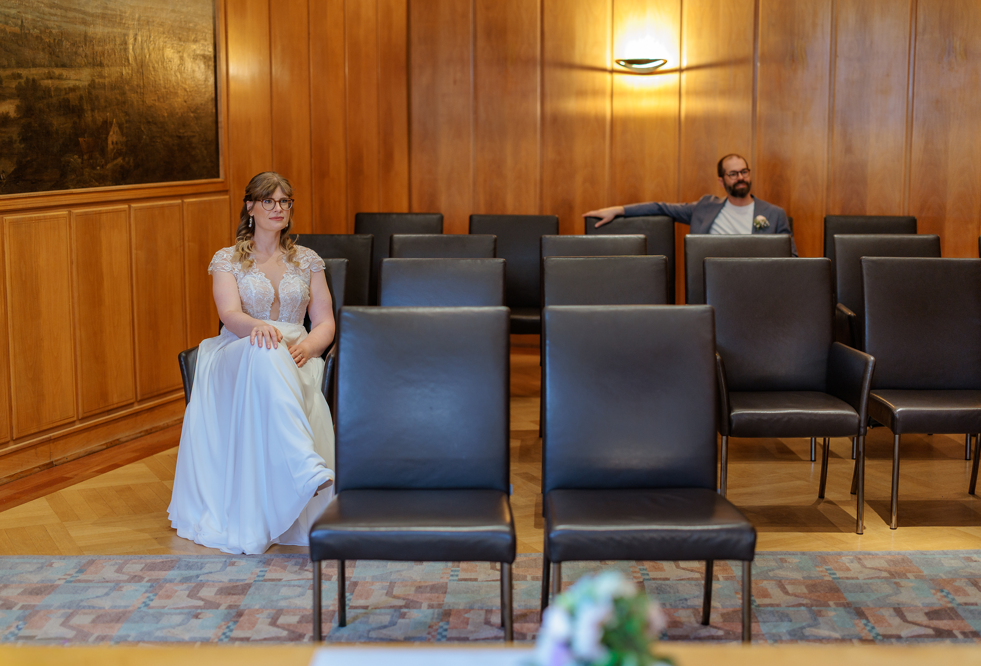 Ein Brautpaar sitzt in einem Saal, in dem die Wandverkleidung aus Holz besteht. Die Braut trägt ein weißes Hochzeitskleid und Brille, während der Bräutigam ein graues Jackett und weiße Kleidung trägt. Es gibt leere Stühle im Vordergrund und im Hintergrund ist eine moderne Wandlampe zu sehen.
