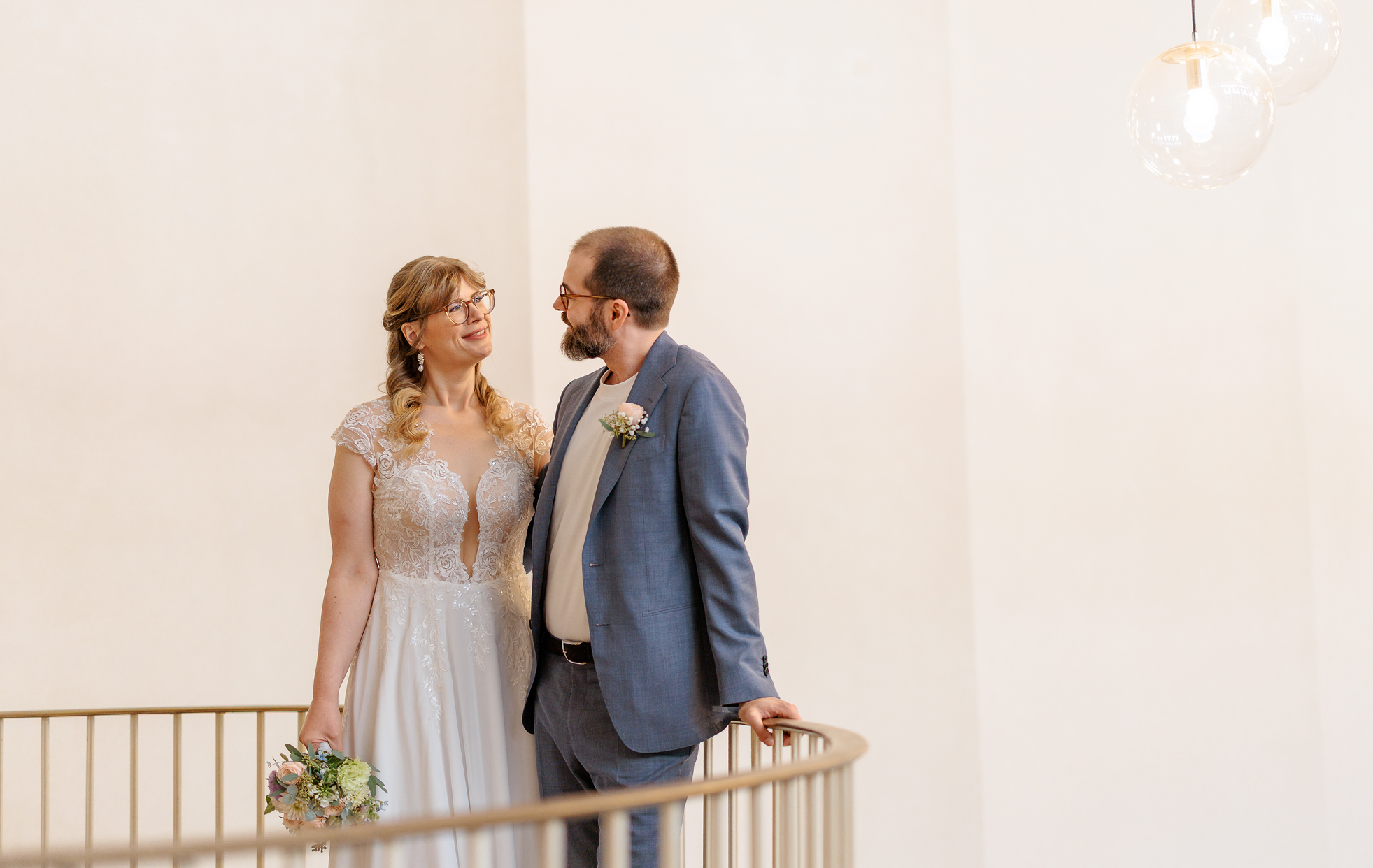 Ein Brautpaar steht auf einer Treppe, die Braut trägt ein weißes Hochzeitskleid mit Spitze, der Bräutigam trägt einen grauen Anzug. Sie schauen sich liebevoll an, die Braut hält einen Brautstrauß in der Hand.
