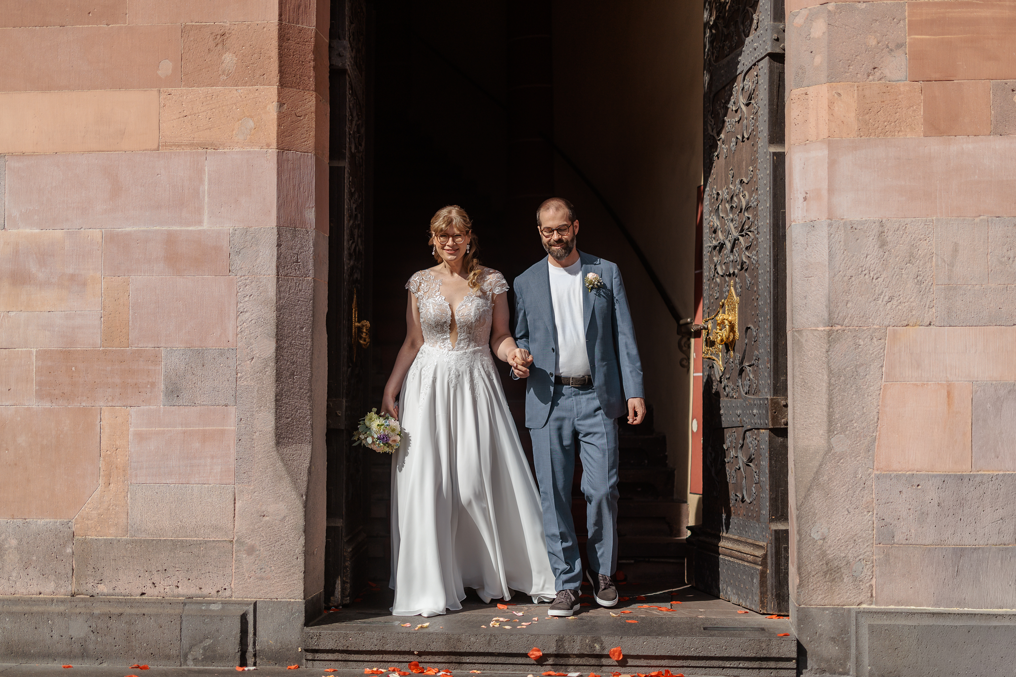 Ein Paar, vermutlich Braut und Bräutigam, halten sich an einer Türöffnung vor einem historischen Gebäude. Die Braut trägt ein weißes Hochzeitskleid mit Spitze und hält einen Blumenstrauß, während der Bräutigam einen Anzug trägt und eine Blume am Revers hat. Sie lachen und gehen gemeinsam hinaus, während Blütenblätter auf dem Boden liegen.