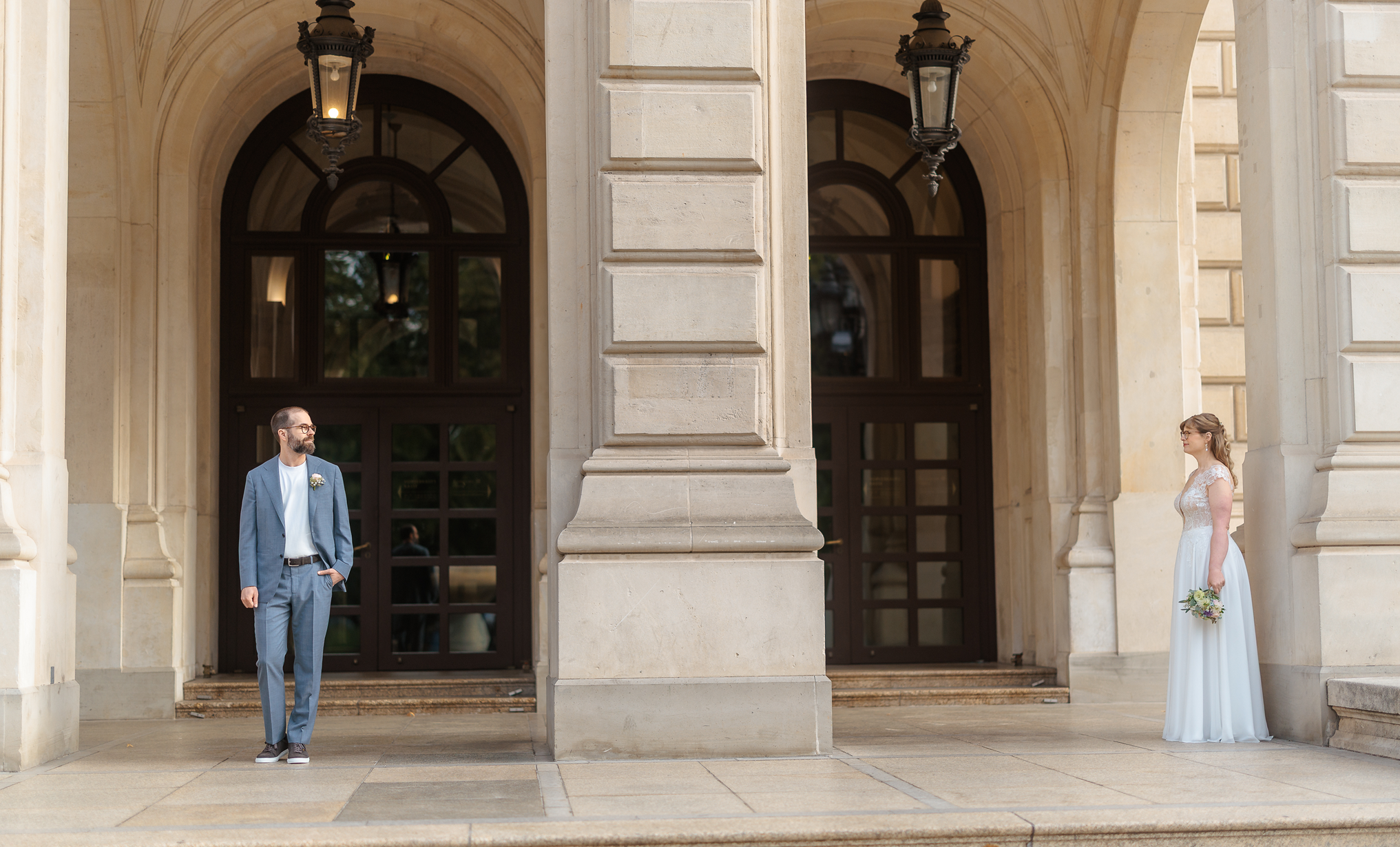 Ein Bräutigam im grauen Anzug steht vor der Alten Oper Frankfurt, während seine Braut im weißen Brautkleid auf der rechten Seite ihren Brautstrauß trägt und in die Ferne blickt.
