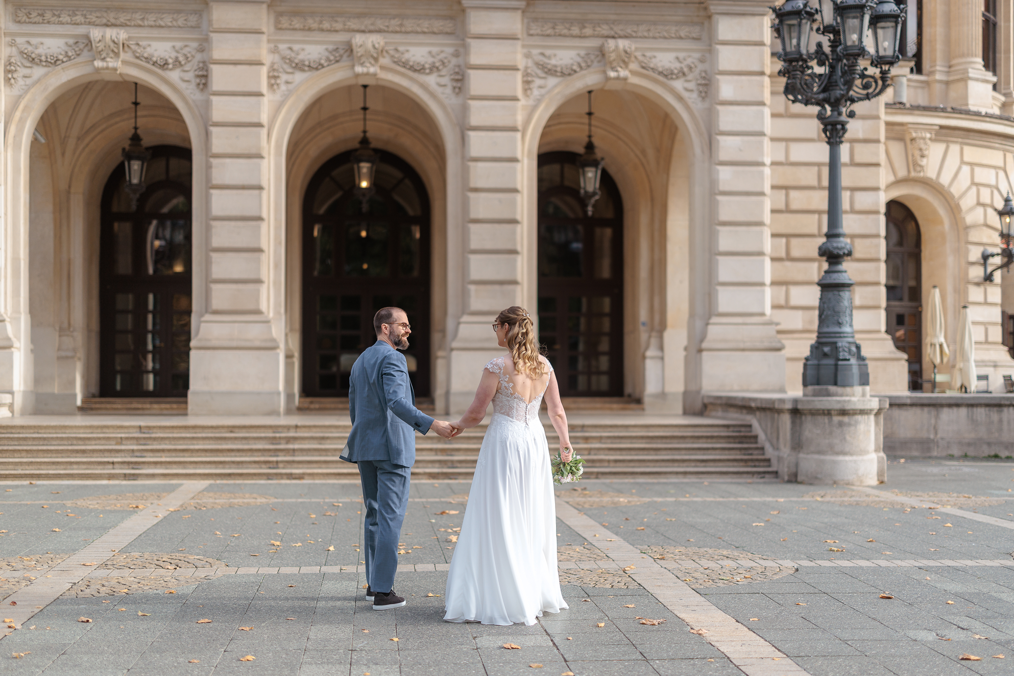 Ein Brautpaar hält sich an Händen vor der Alten Oper in Frankfurt im Vordergrund liegen herabfallende Herbstblätter auf den Pflastersteinen.