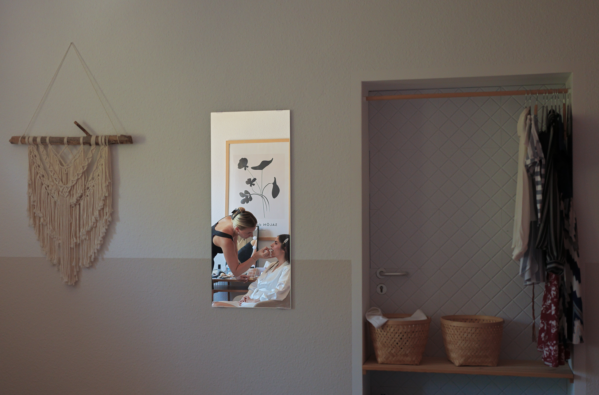 Ein Friseur macht einer Braut Make-up, während sie im Spiegel schaut, in einem Raum mit weißer Wand, dekoriert mit einem makrame-Wandbehang, und einem offenen Kleiderschrank mit Kleidungsstücken.