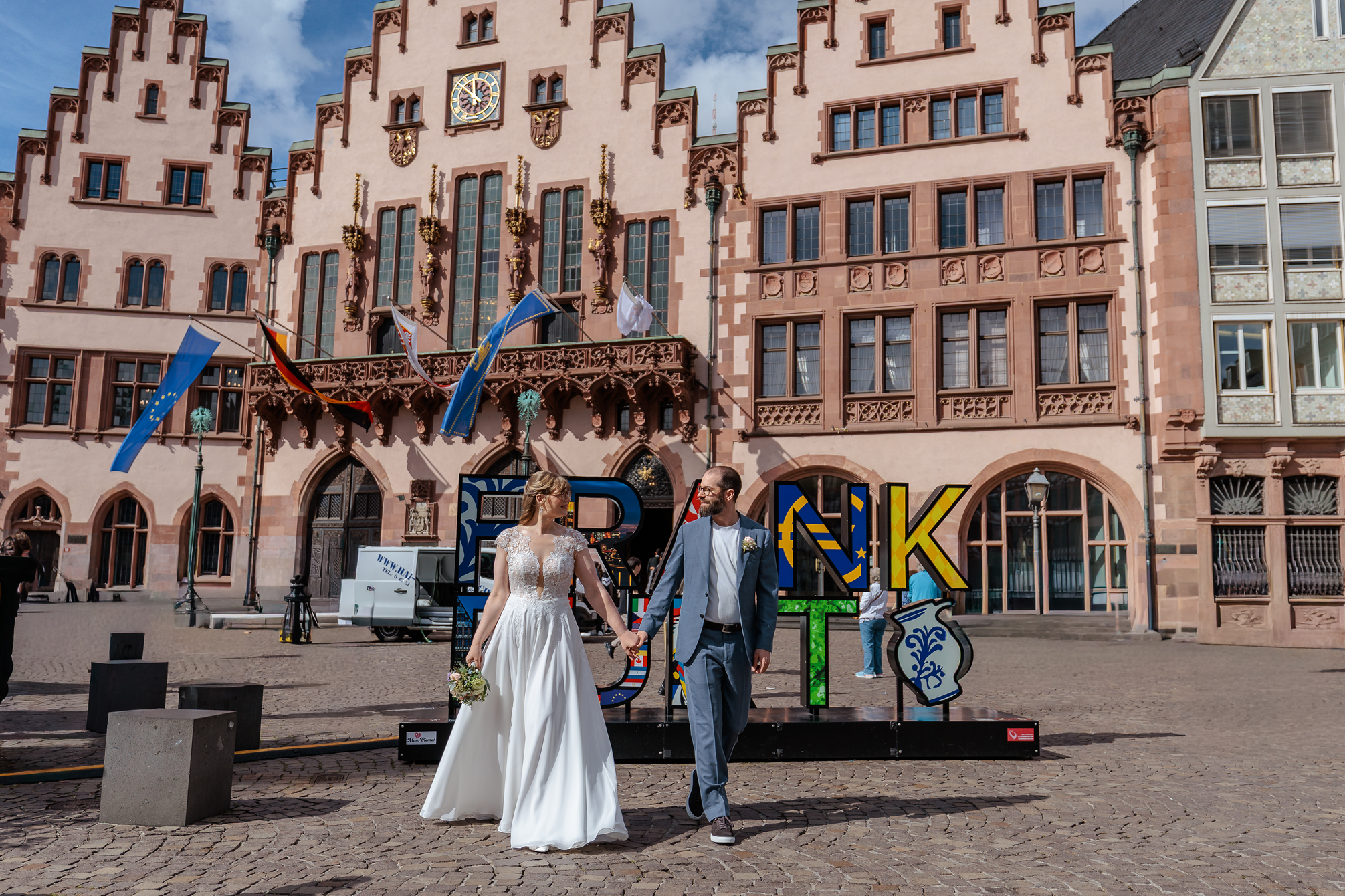 Ein frisch vermähltes Paar, eine Frau in einem weißen Brautkleid und ein Mann in einem grauen Anzug, hält Händchen vor einem historischen Rathaus in Deutschland. Im Hintergrund sind Deutschland- und EU-Flaggen sowie bunte Buchstaben und Dekorationen für einen Festakt sichtbar.