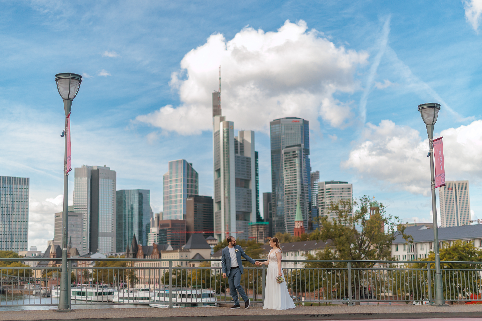 Ein Paar, vermutlich Braut und Bräutigam, hält sich an einem Fluss mit einer Stadt im Hintergrund, darunter Hochhäuser und Wolkenkratzer bei sonnigem Himmel.