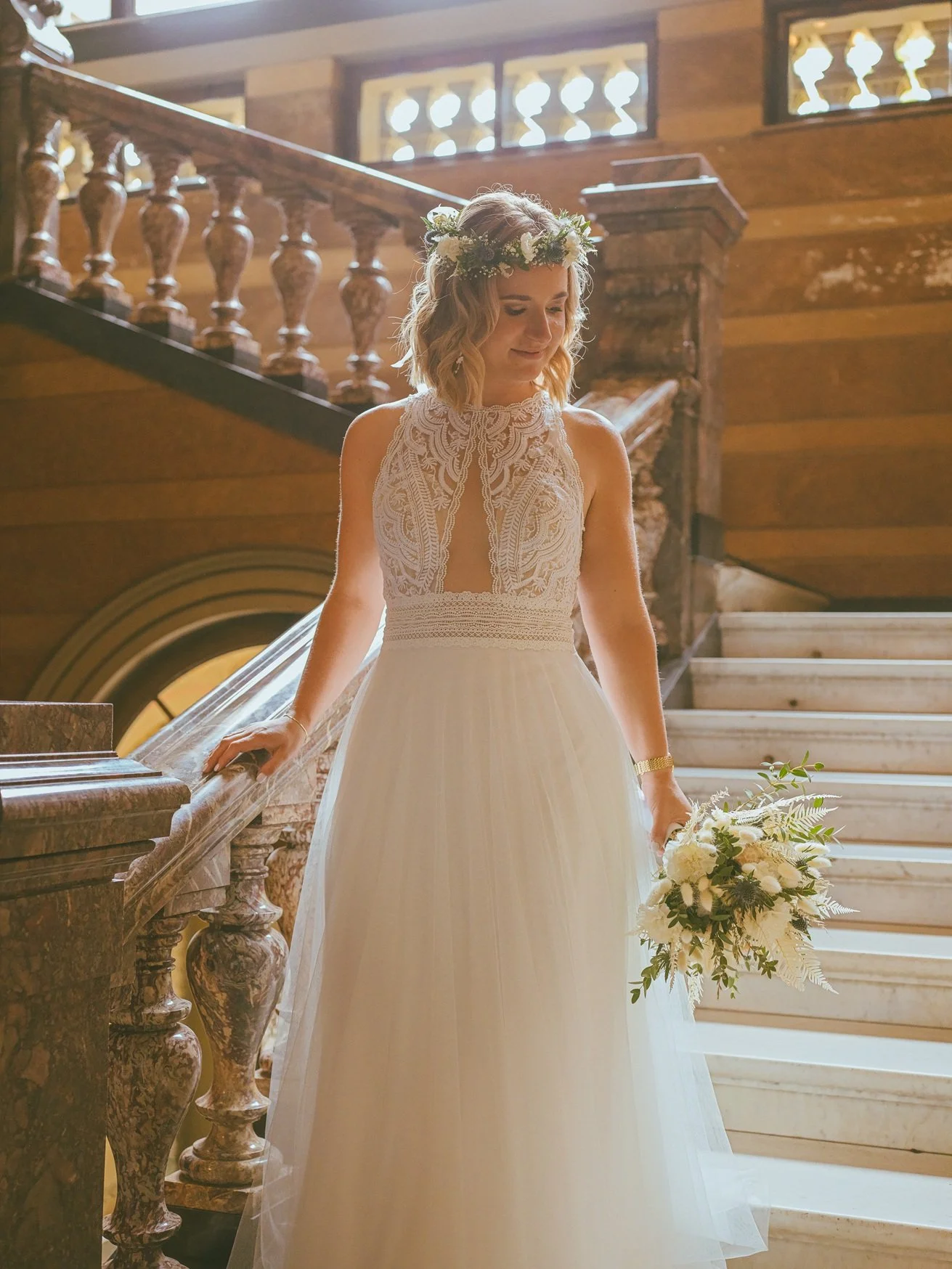 Eine Frau in einem weißen Brautkleid mit Blumenkranz auf dem Kopf steht auf einer Treppe im Standesamt der Villa Böhm in Neustadt und hält einen Blumenstrauß in der Hand.