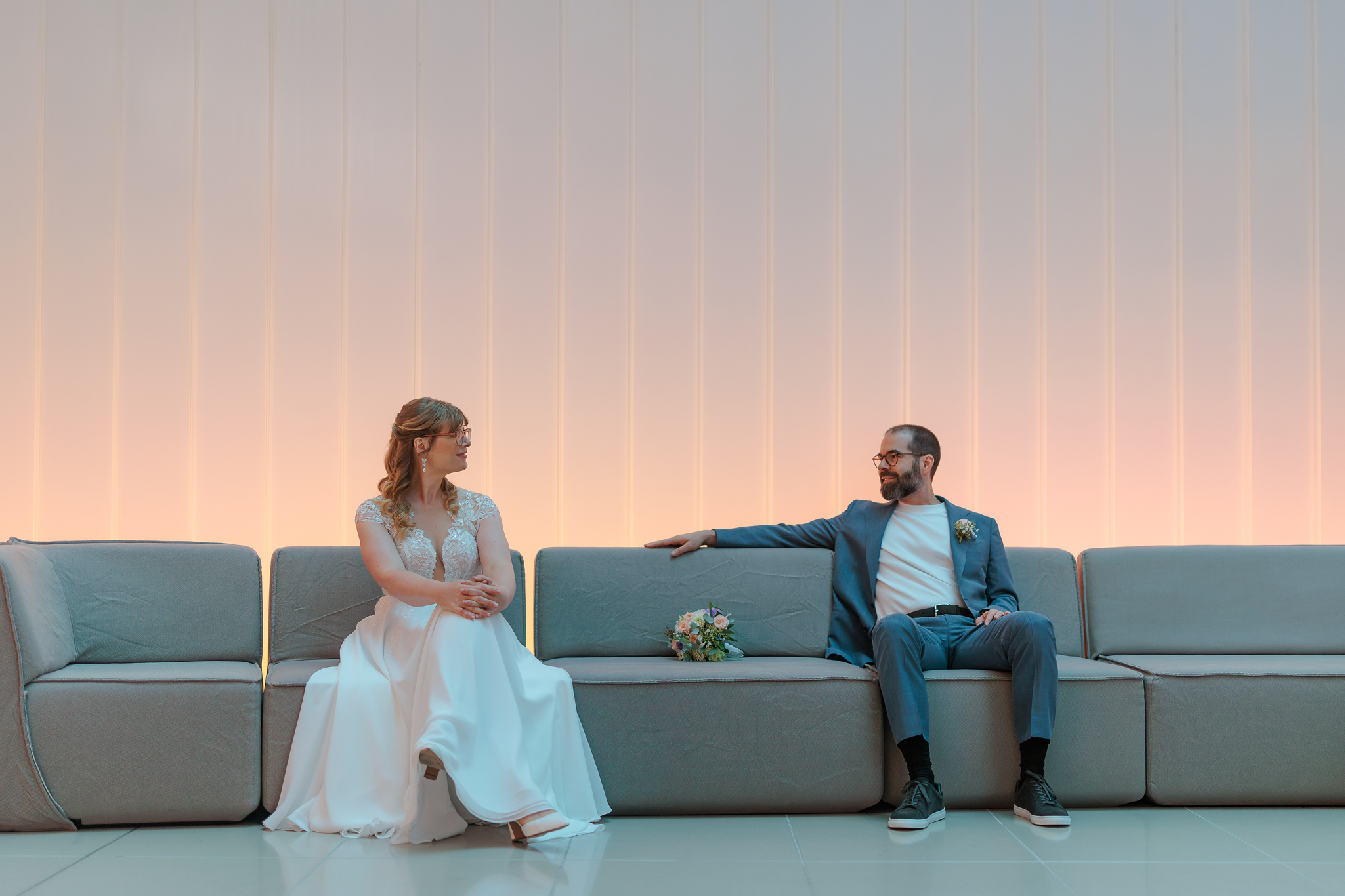 Ein Brautpaar sitzt auf einer Couch im Foyer des Eurotheum Frankfurt, sie trägt ein Hochzeitskleid mit Spitzendetail, er trägt einen Anzug ohne Krawatte. Sie schaut sich an, im Hintergrund eine hell erleuchtete Wand.