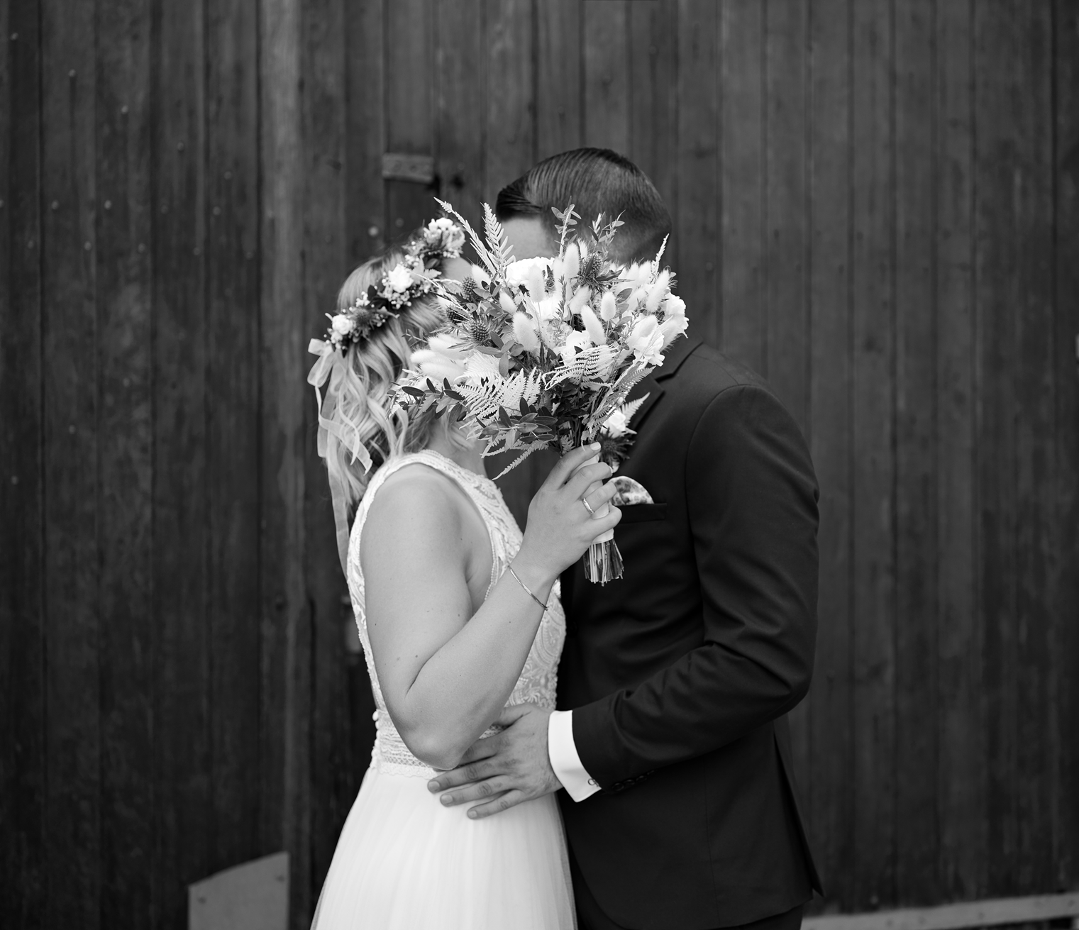 Verliebtes Brautpaar bei ihrer Hochzeit, die Braut mit Blumenkranz auf dem Kopf hält einen Blumenstrauß vor das Gesicht des Bräutigams.
