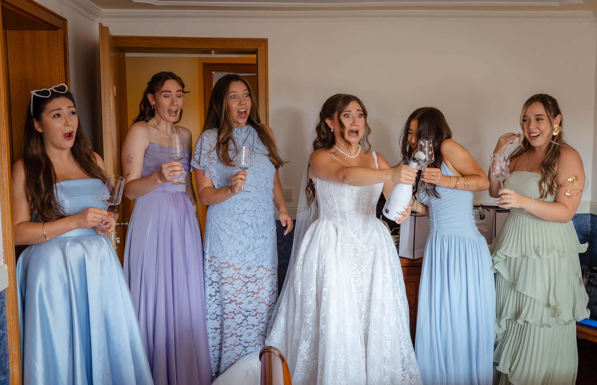 Eine Gruppe von Frauen, wahrscheinlich Brautjungfern, lacht und wirkt überrascht während einer Hochzeitsfeier in einem Wohnzimmer.