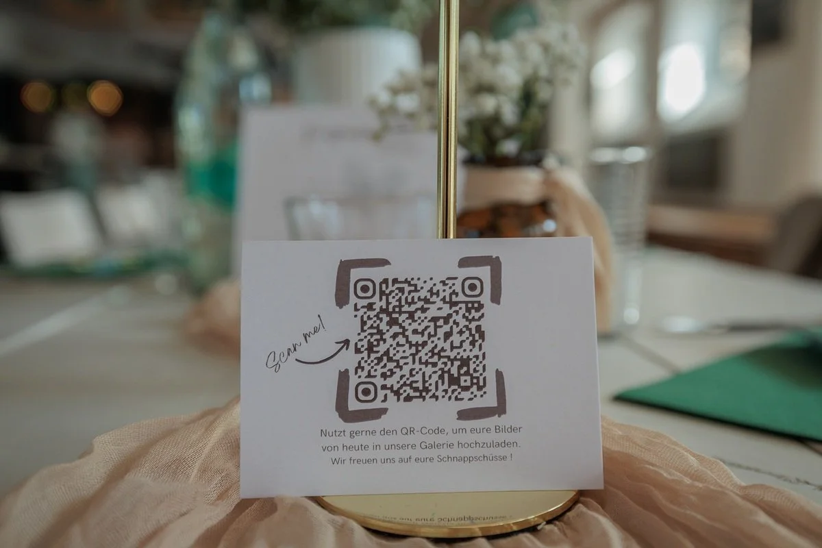 QR-Code auf einer Karte, mit der Aufforderung zum Hochladen von Bildern in einer Galerie, in der Hochzeitslocation Klosterscheune Wehrheim, mit Blumentopf im Hintergrund.