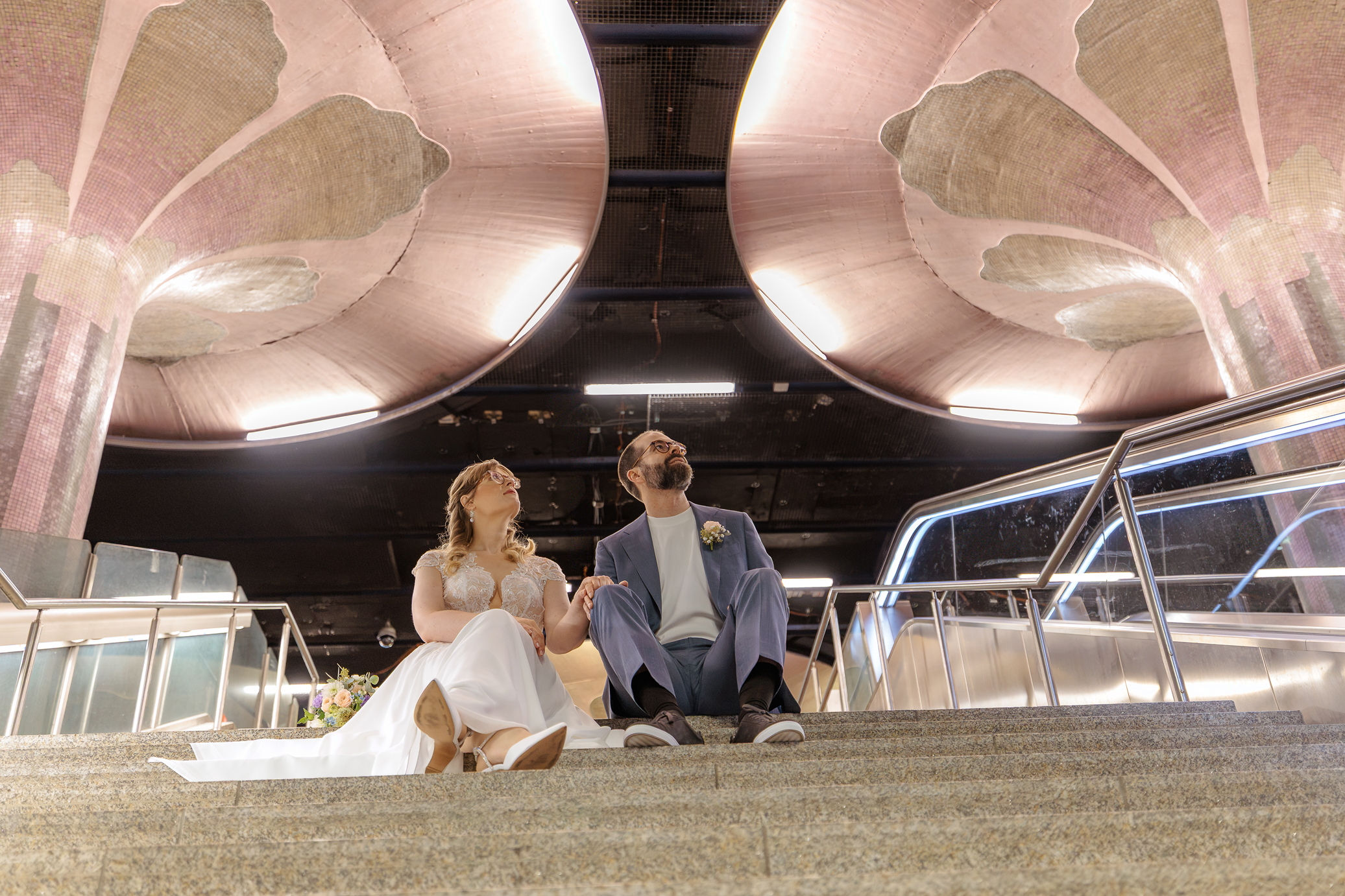 Ein Brautpaar sitzt auf Treppen im U-Bahnhof Frankfurt Westend, verliebt und entspannt, während sie ihre Hochzeit feiern.