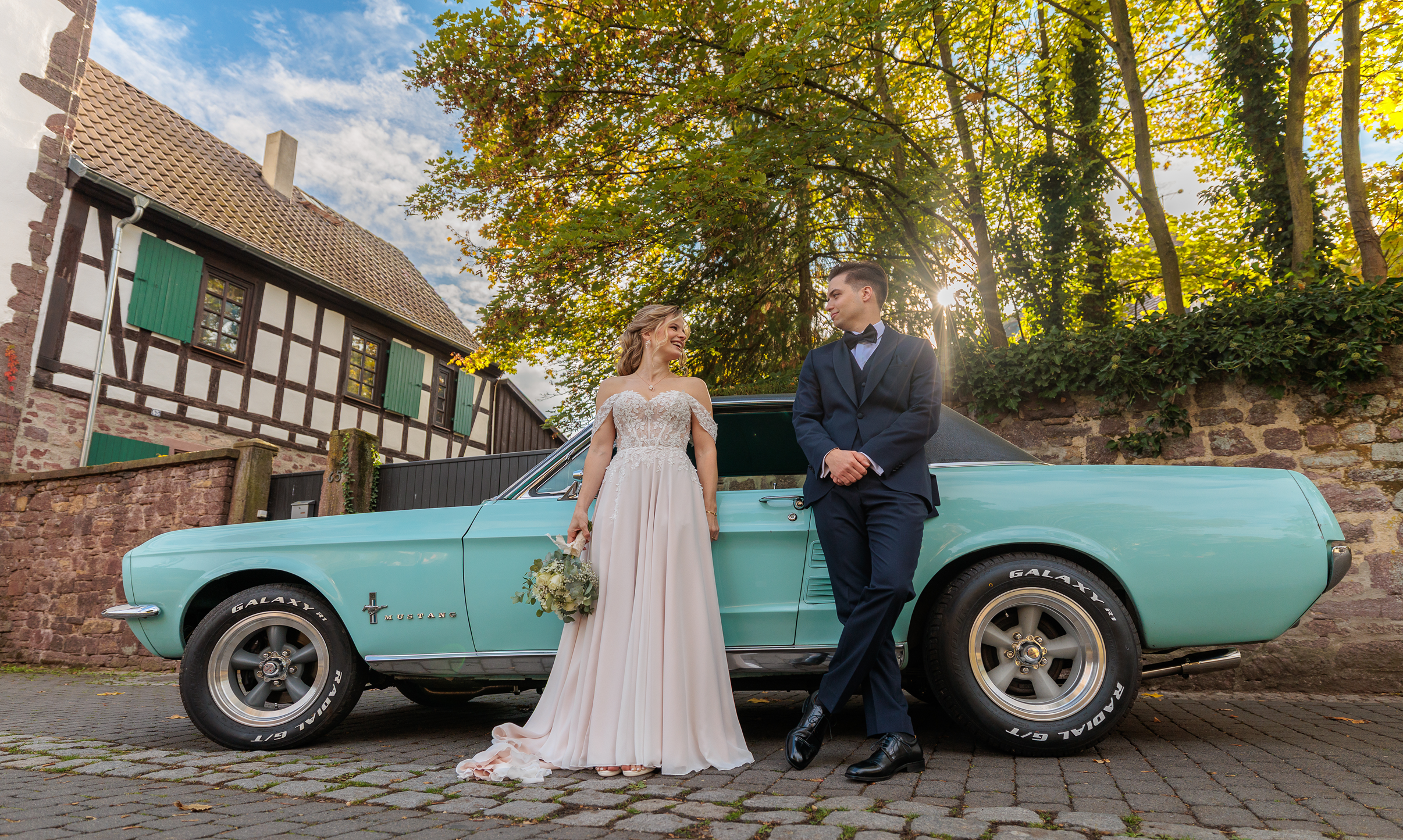 Ein Brautpaar steht neben einem alten blauen Mustang-Auto vor einem historischen Fachwerkhaus gegenüber der Burg Hayn, die Sonne scheint durch die Bäume im Hintergrund.