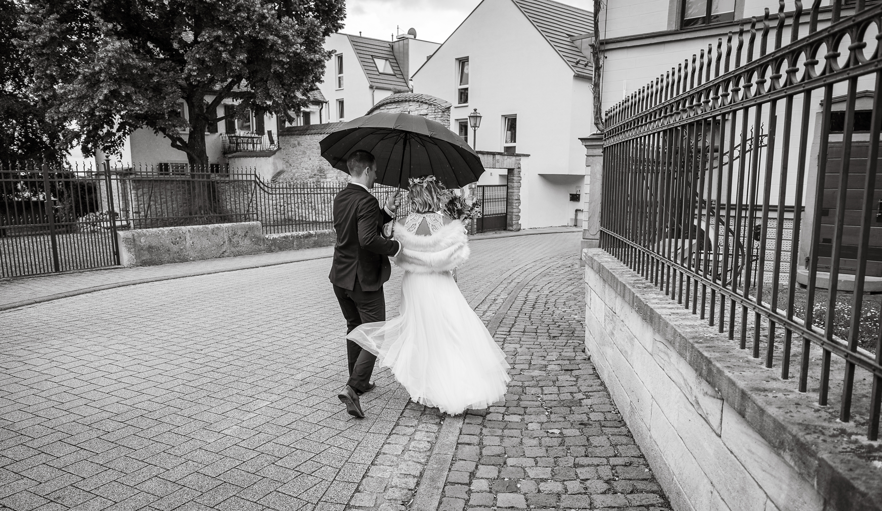 Ein Brautpaar geht bei Regen unter einem schwarzen Regenschirm. Die Braut trägt ein langes weißes Kleid und eine Blumenkrone, der Bräutigam einen schwarzen Anzug. Sie gehen auf einer gepflasterten Straße in einer Stadt.