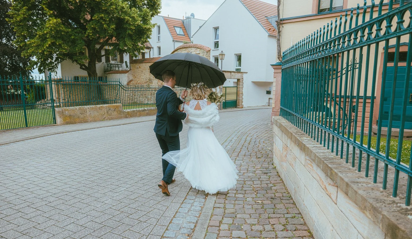 Ein Mann und eine Frau bei einer Hochzeit, der Mann hält einen schwarzen Regenschirm über die Braut, während sie gemeinsam gehen, auf einer gepflasterten Straße, bei leichtem Regen.
