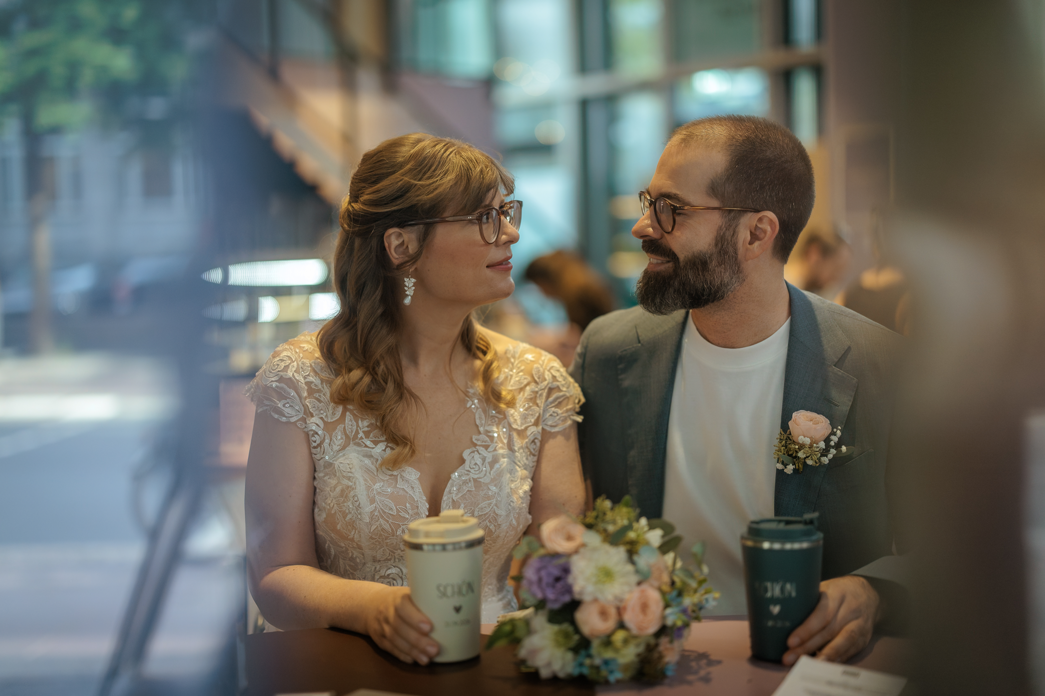 Ein Paar, vermutlich Braut und Bräutigam, sitzen im Café und betrachten sich liebevoll. Die Frau trägt ein weißes Spitzenkleid und Brillengläser, hält einen Kaffeebecher und hat einen Blumenstrauß vor sich. Der Mann trägt einen grauen Anzug, Brille und hat einen Bart. Im Hintergrund sind große Fenster und andere Gäste zu sehen.