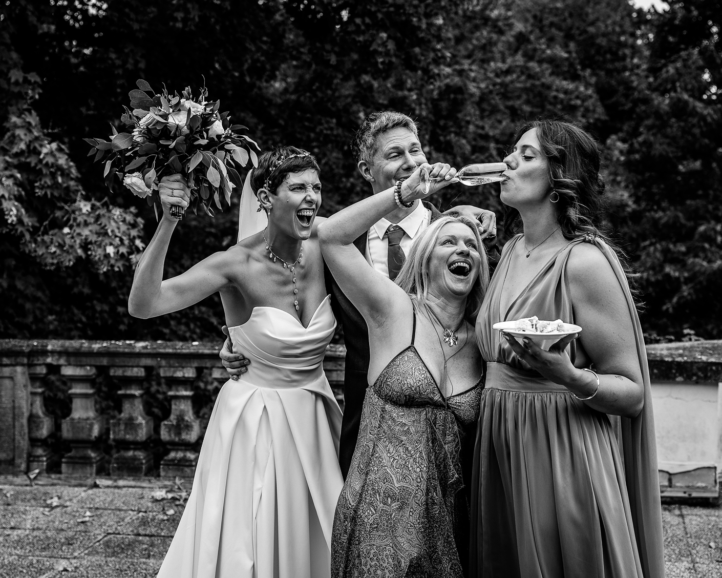 Junge Frau in Brautkleid bei Hochzeit, umgeben von drei Frauen, eine in Brautkleid, eine in Abendkleid, alle lachen, eine hält einen Blumenstrauß, andere trinken Sekt, outdoor im Park des Dolce Bad Nauheim, schwarz-weiß