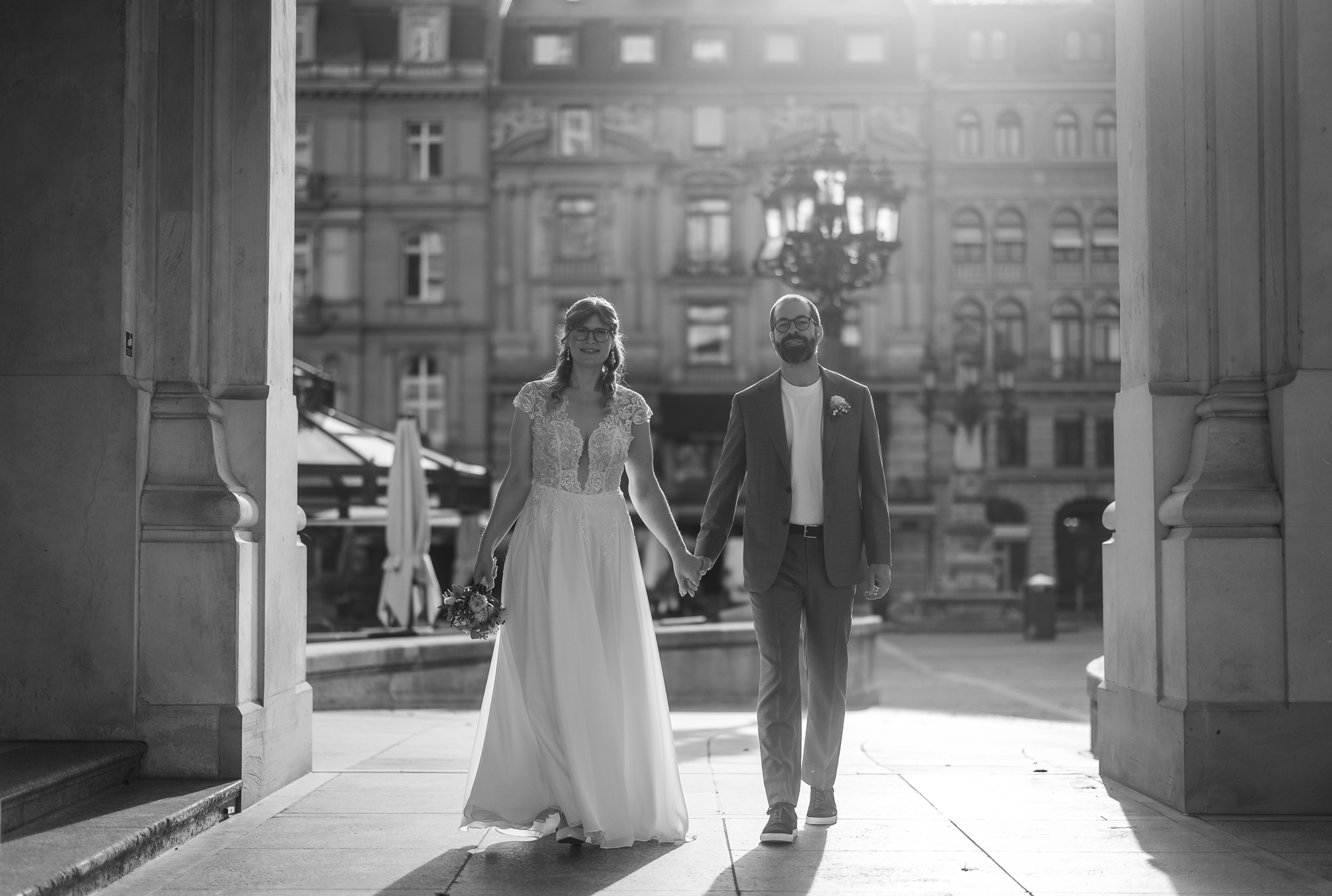 Braut und Bräutigam gehen Hand in Hand durch eine Säulenöffnung der Alten Oper Frankfurt, beide lächeln, es ist ein sonniger Tag, im Hintergrund alte Gebäude und Straßenlaternen, schwarz-weiß Foto.