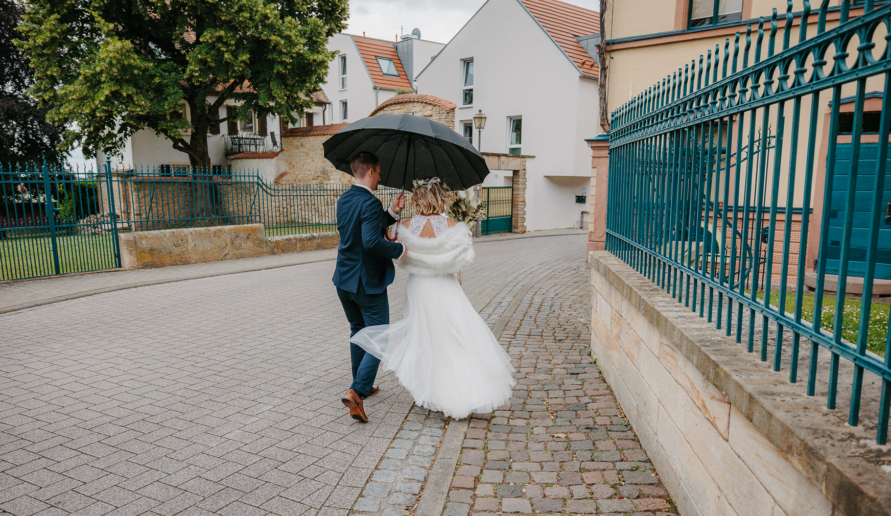 Ein Brautpaar geht an einem regnerischen Tag zusammen unter einem schwarzen Regenschirm, die Braut trägt ein weißes Hochzeitskleid mit Schleier, der Bräutigam trägt einen dunklen Anzug, während sie auf einem gepflasterten Gehweg neben einer Mauer und einem grünen Zaun gehen.
