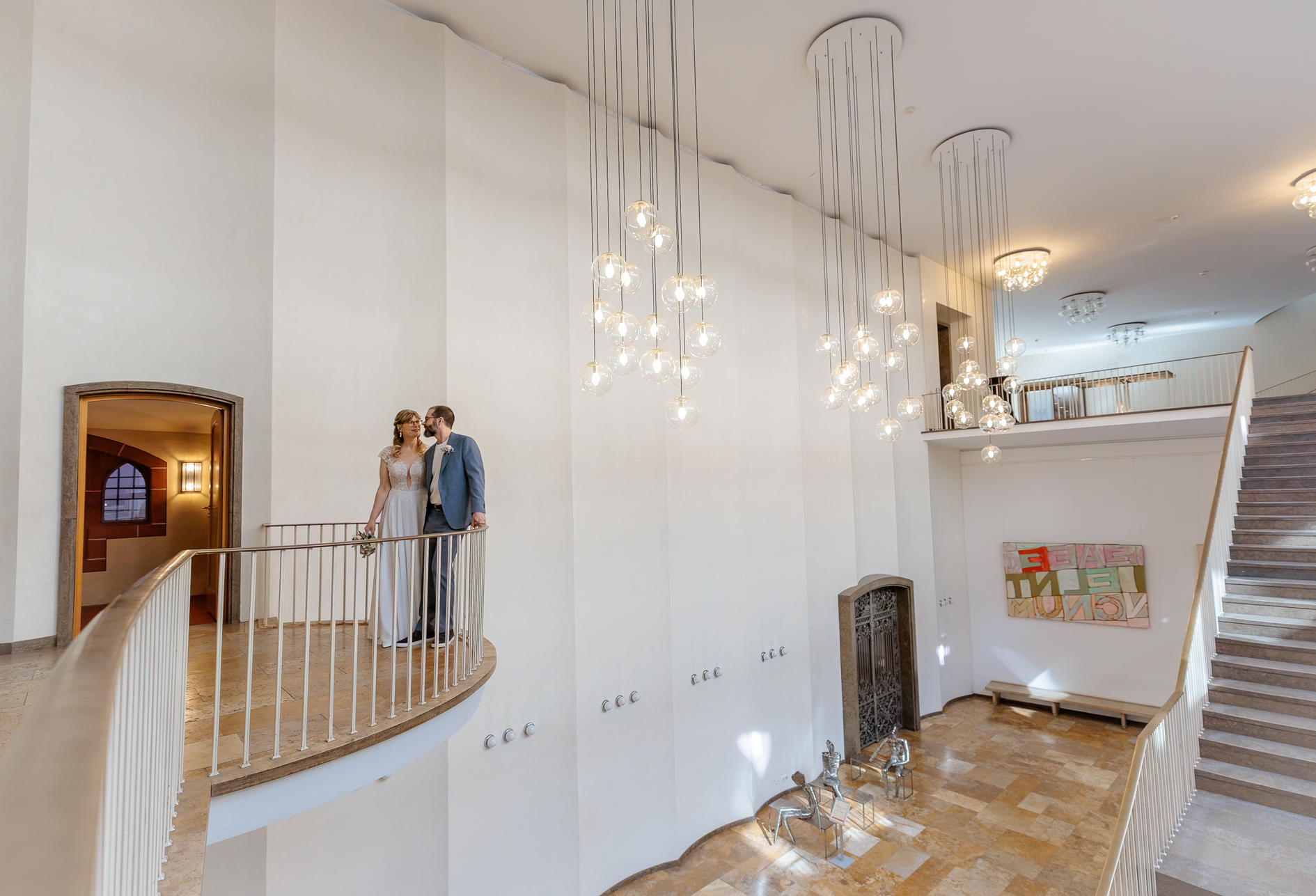 Ein Brautpaar steht auf einer weißen Galerie im Innenraum eines Hauses, umgeben von modernen Lichtern und Kunstwerken.