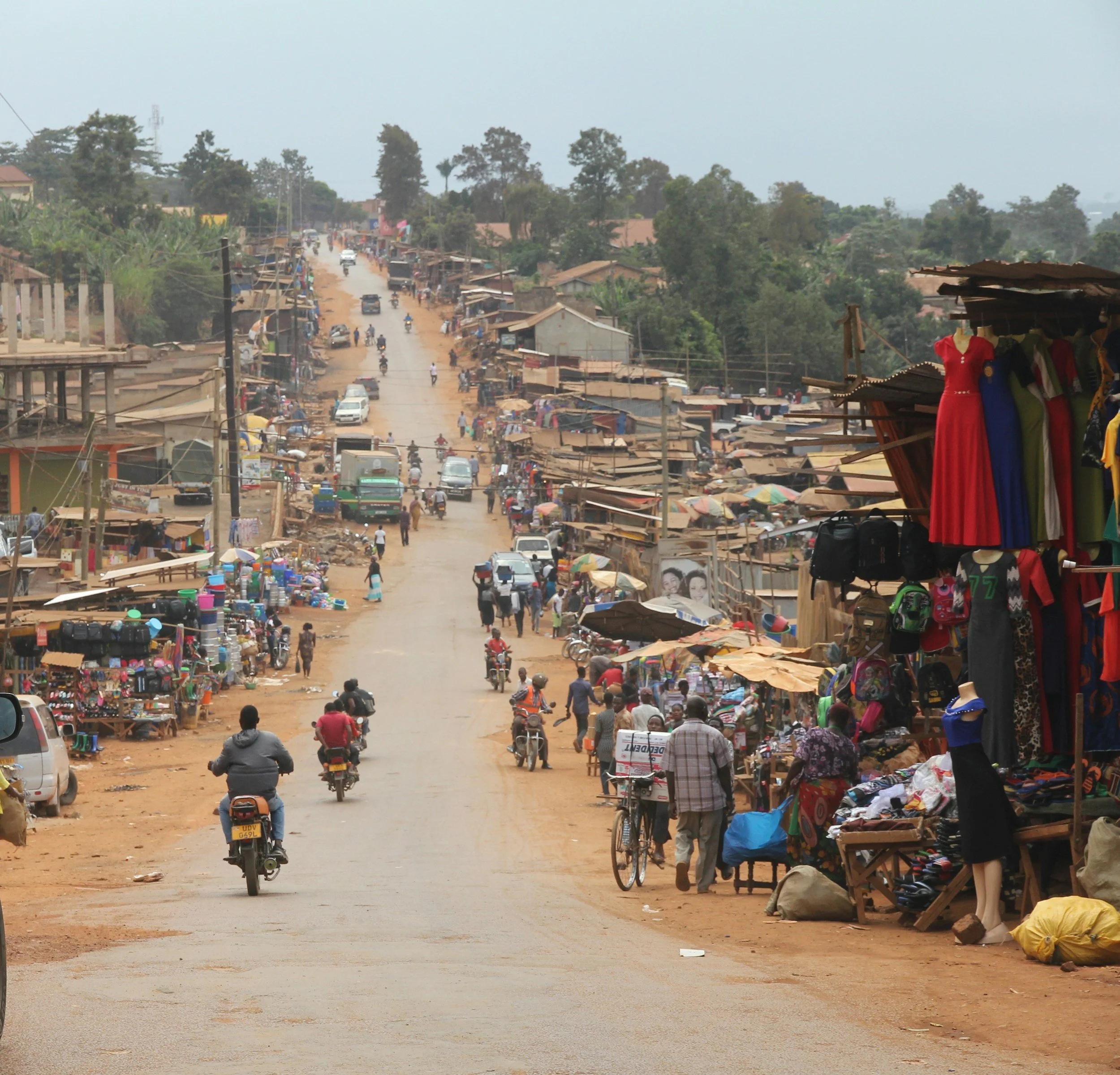Bustling street market scene with people, motorcycles, and vendor stalls displaying clothing and various goods.